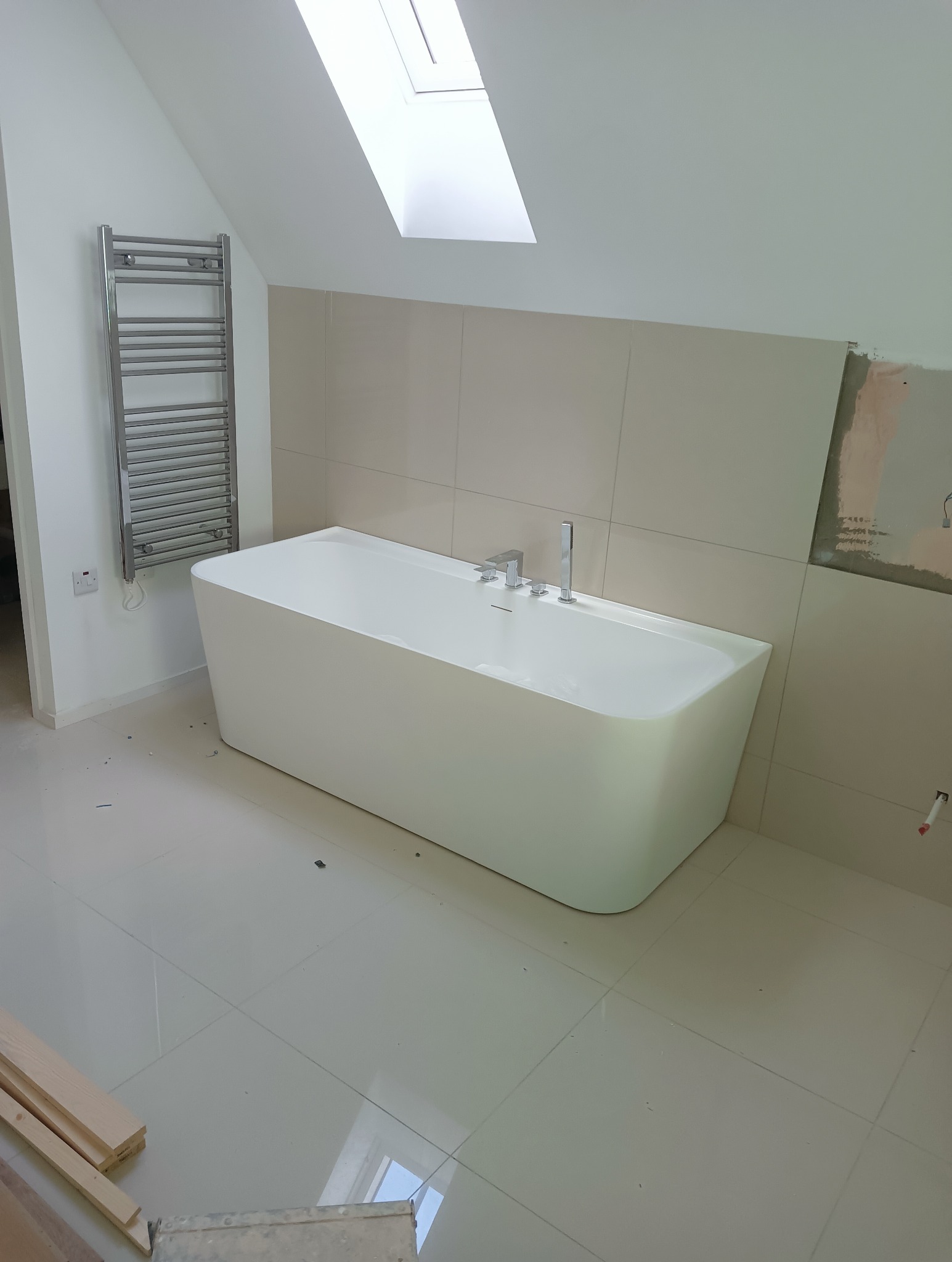 Nowoczesna, biała wanna wolnostojąca w jasnej łazience z oknem dachowym, grzejnikiem drabinkowym i beżowymi płytkami ściennymi. Częściowo wykończona ściana.