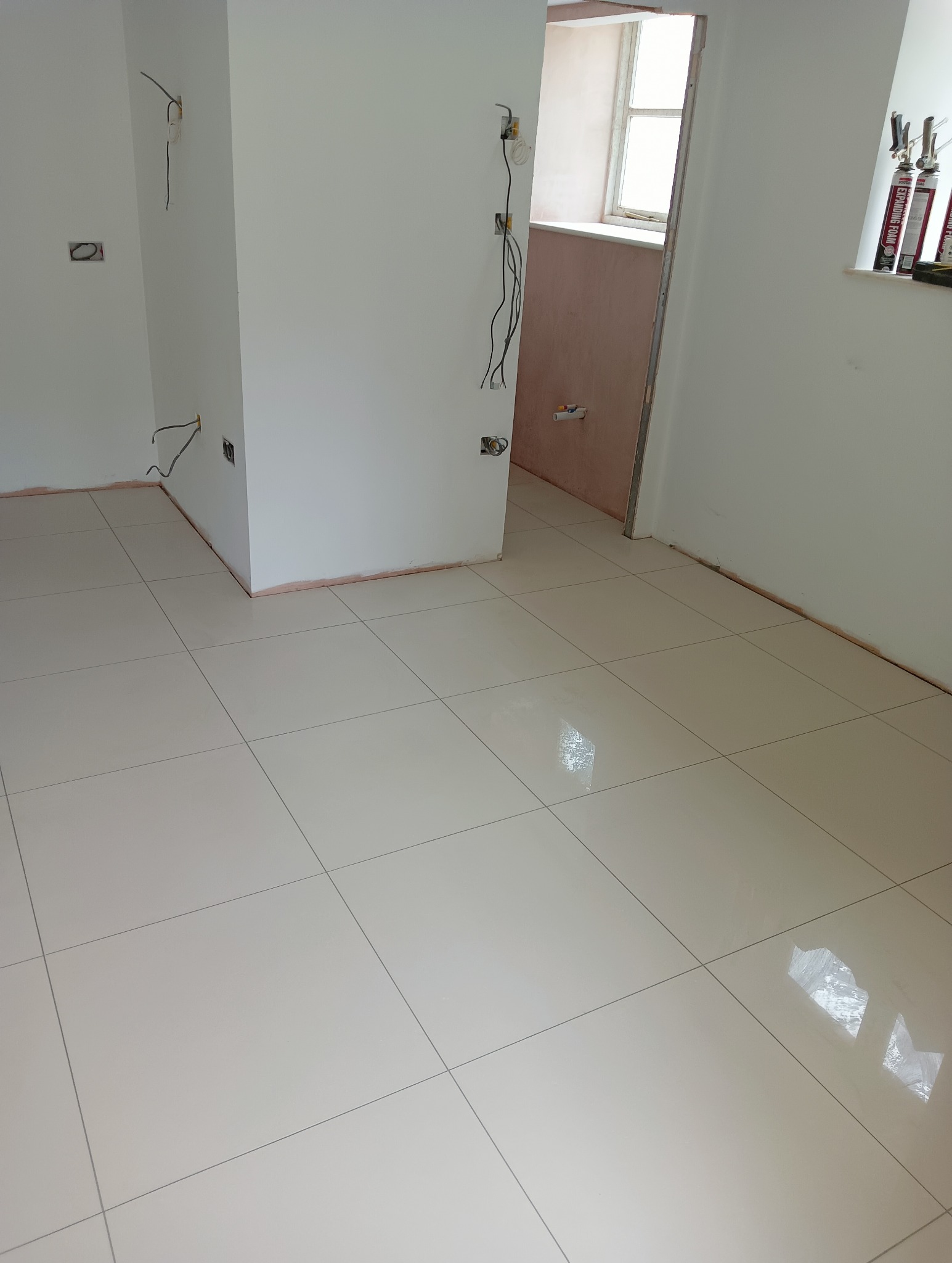 Jasne, błyszczące płytki podłogowe ułożone w nowym pomieszczeniu. Widoczne przygotowane ściany z instalacją elektryczną i puszki z pianką montażową na parapecie.