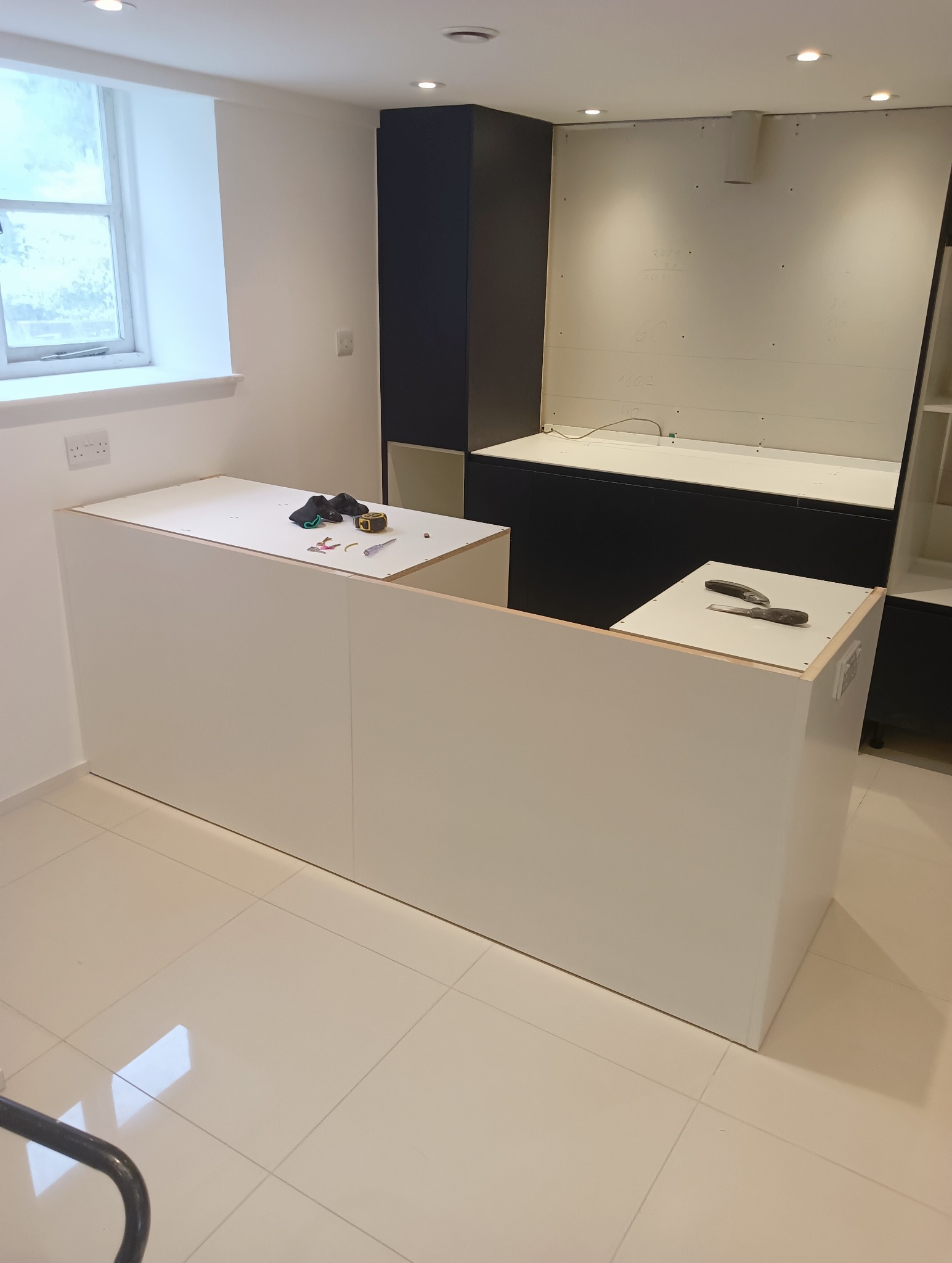 Montaż białej zabudowy kuchennej z ciemnymi elementami. Widoczne narzędzia na blacie, okno w tle i oświetlenie sufitowe.