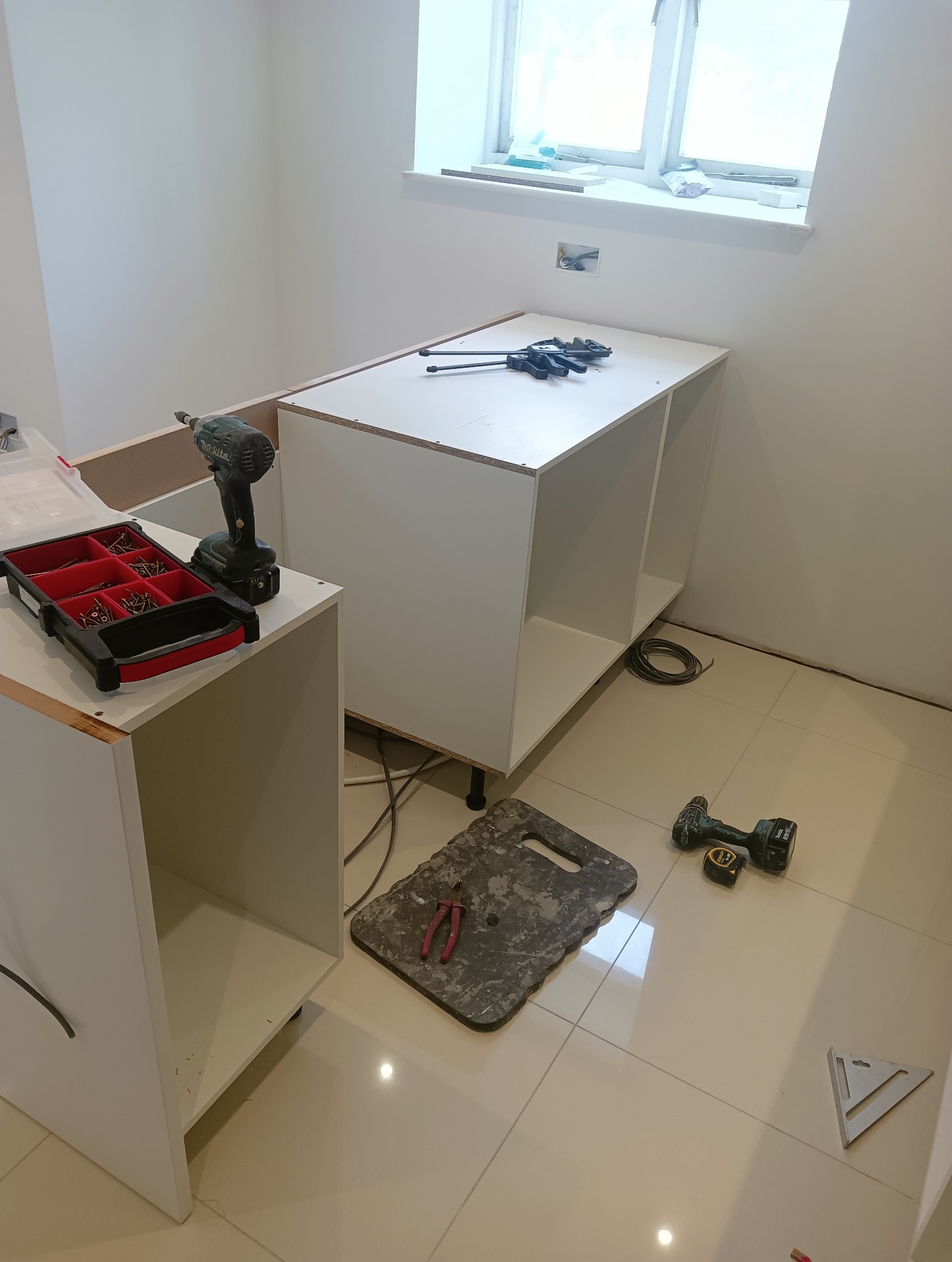 Montaż białych szafek kuchennych pod oknem. Widoczne narzędzia: wiertarka, skrzynka ze śrubami, miarka, kątownik, klamry stolarskie. Etap montażu, surowe wnętrze.