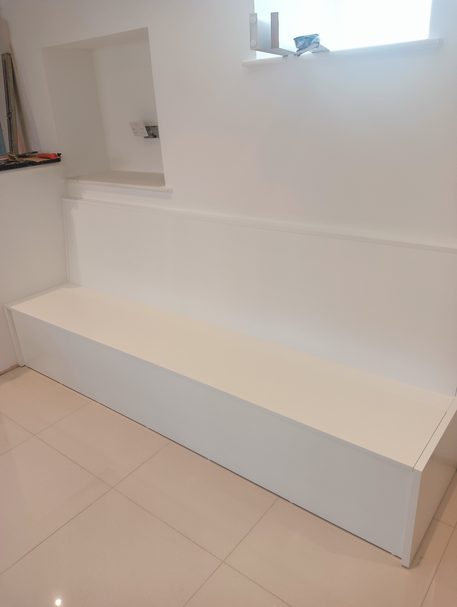 Biała, wnękowa ławka w minimalistycznym wnętrzu. Świeżo pomalowane ściany, jasne płytki podłogowe. Widoczne gniazdka elektryczne w ścianie i narzędzia malarskie.