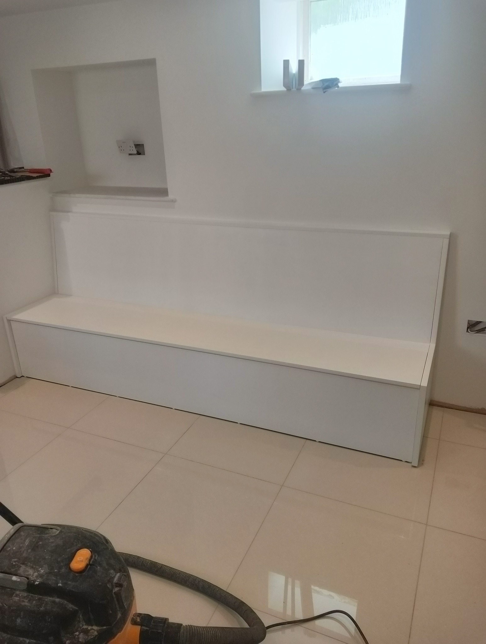 Biała zabudowa wnęki z siedziskiem pod oknem, świeżo po montażu. Widoczne gniazdka elektryczne, płytki podłogowe i odkurzacz.