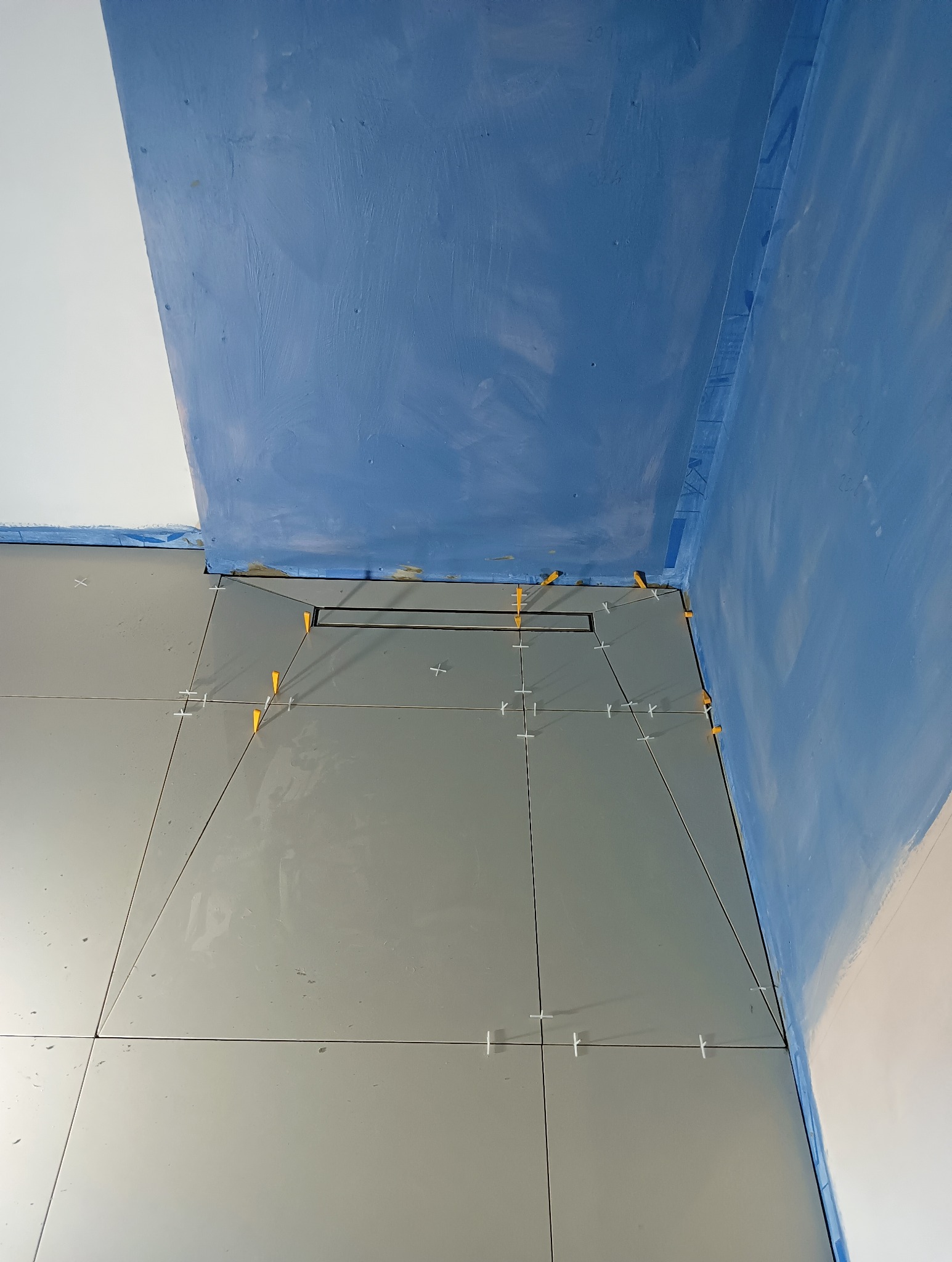 Szare płytki podłogowe w łazience, ułożone z zachowaniem spadku w kierunku odpływu liniowego, z użyciem żółtych i białych krzyżyków dystansowych.