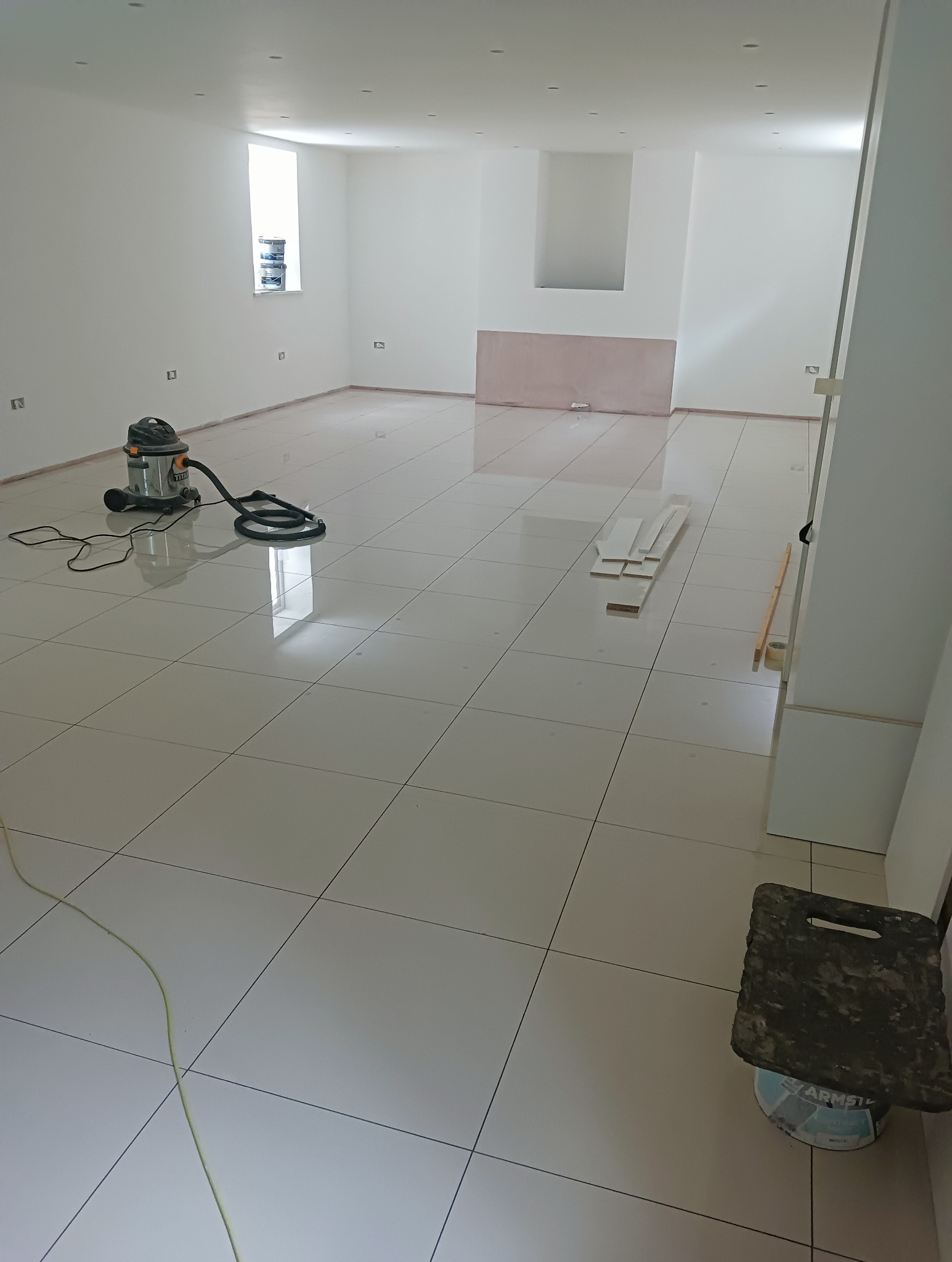 Białe, błyszczące płytki podłogowe w pustym, odnowionym pomieszczeniu. Widoczny odkurzacz, przymocowane listwy przypodłogowe i puszki farby. Minimalistyczne wnętrze.