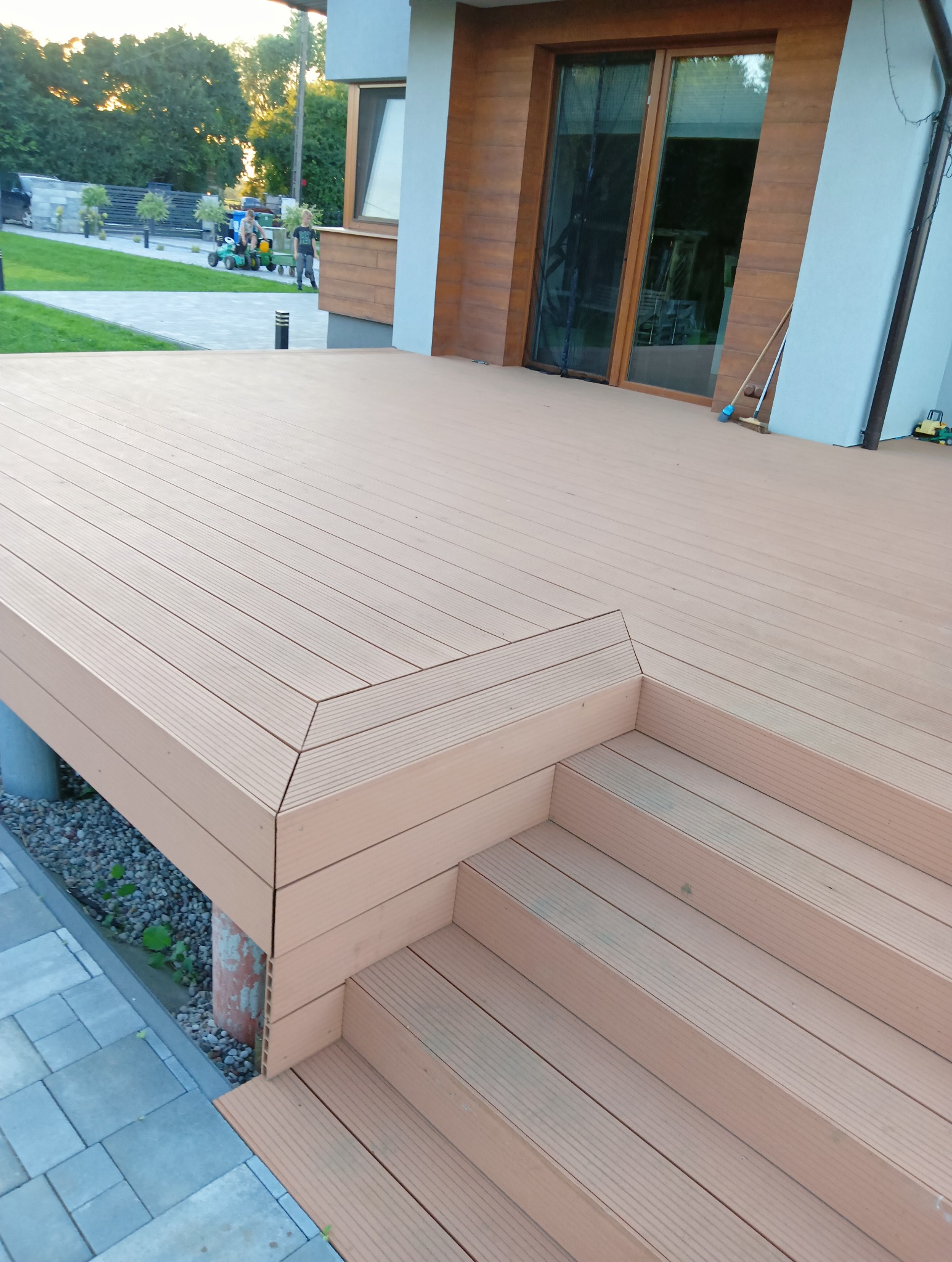 Nowy taras z kompozytu, ze schodami, w odcieniach brązu, przylegający do nowoczesnego domu z dużymi oknami. Widok na zadbany trawnik i bawiące się dzieci.