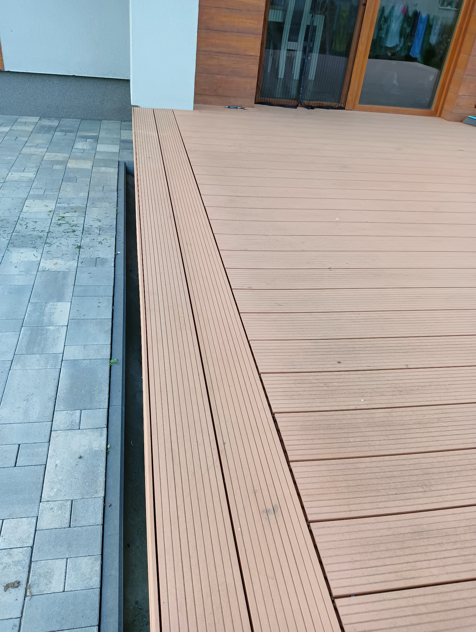 Kompozytowa deska tarasowa w odcieniach brązu, ułożona przy szarej kostce brukowej. Widoczna faktura desek i ich regularny układ. Tło stanowi jasna elewacja budynku i drewniane drzwi.