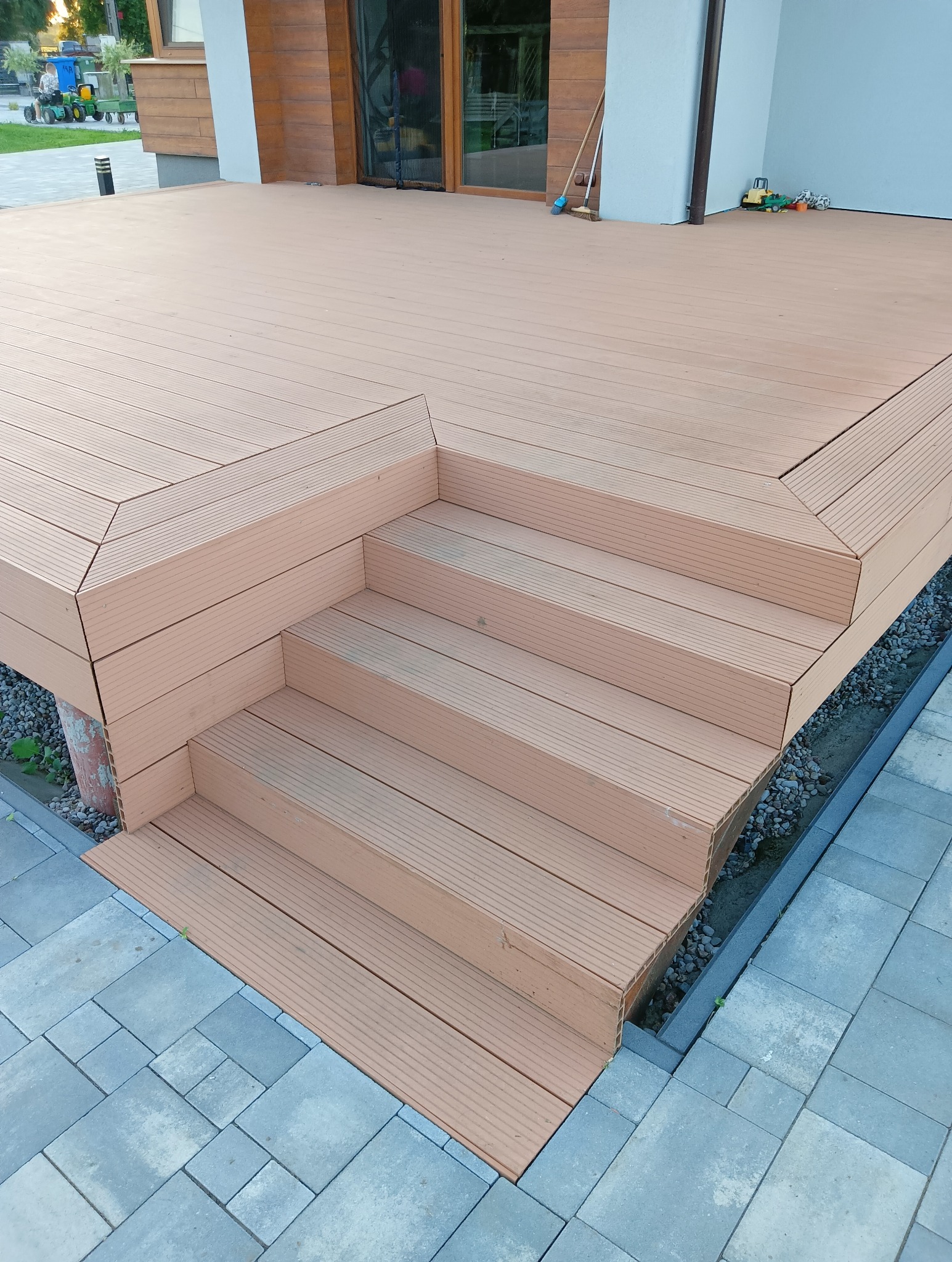 Nowy, brązowy taras kompozytowy z trzema schodami, wykończony szarą kostką brukową. Widoczne detale łączeń desek i krawędzi. W tle dom z drewnianą elewacją.