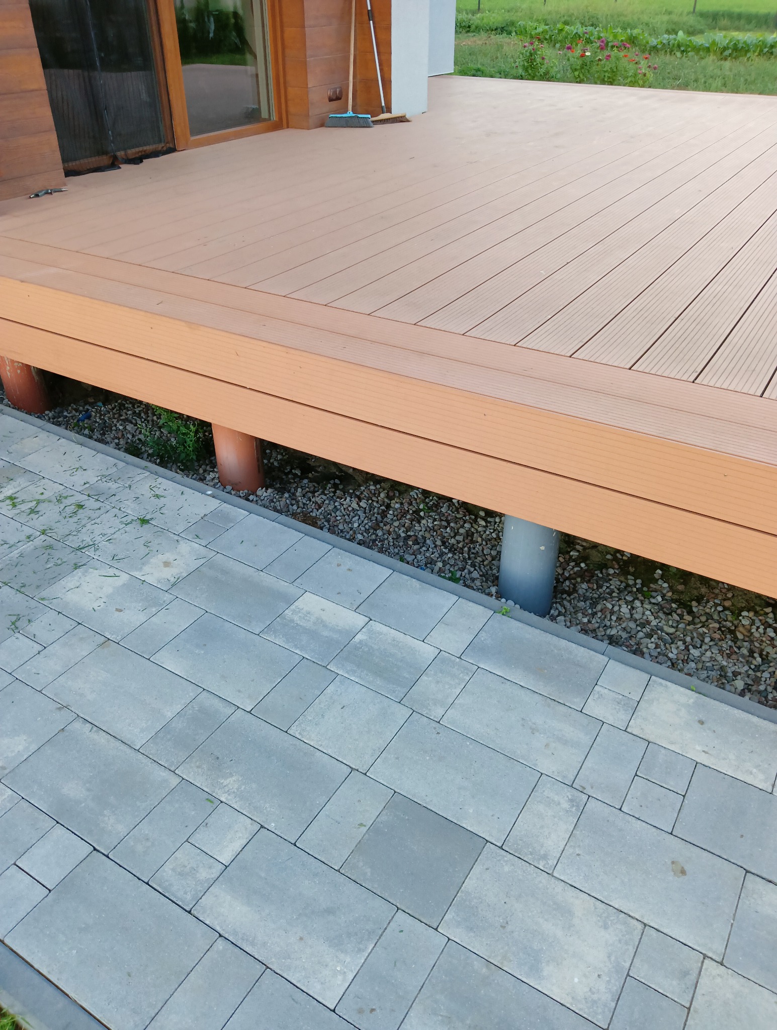 Wykończony taras z kompozytowych desek, wsparty na rurach, widok z boku na szarą kostkę brukową i rabatę z kamykami. Widoczne narzędzia do sprzątania.