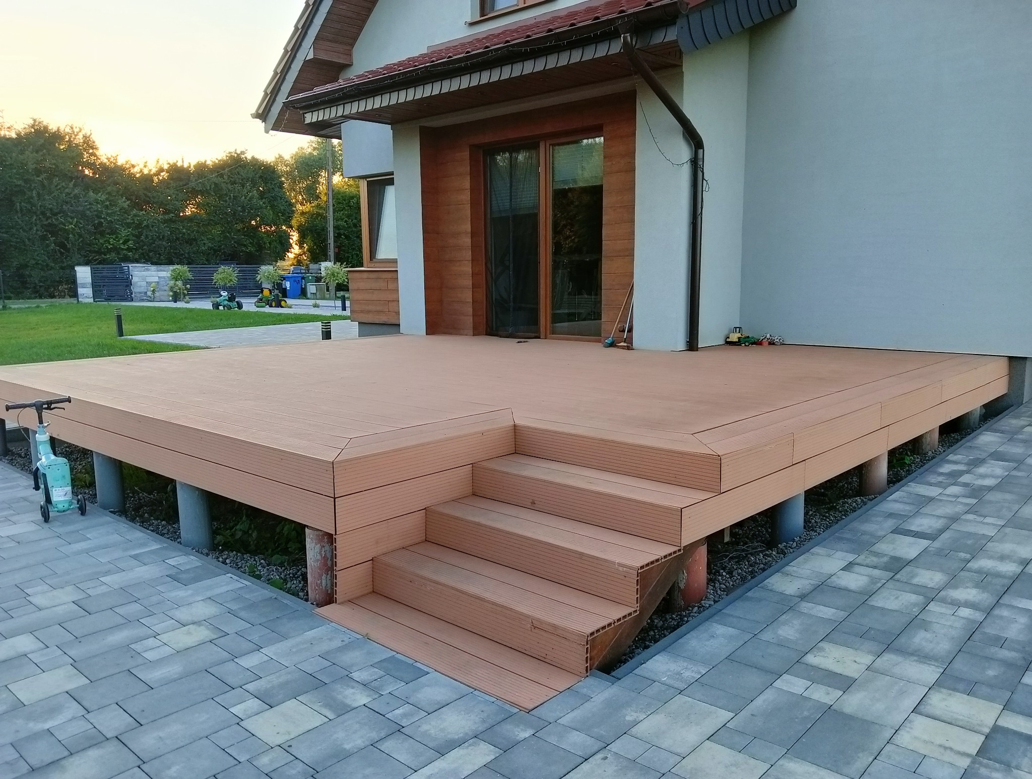 Nowy taras z kompozytowych desek w kolorze drewna, z niskimi schodami, na tle domu z drewnianą elewacją i kostki brukowej. Widok na ogród.