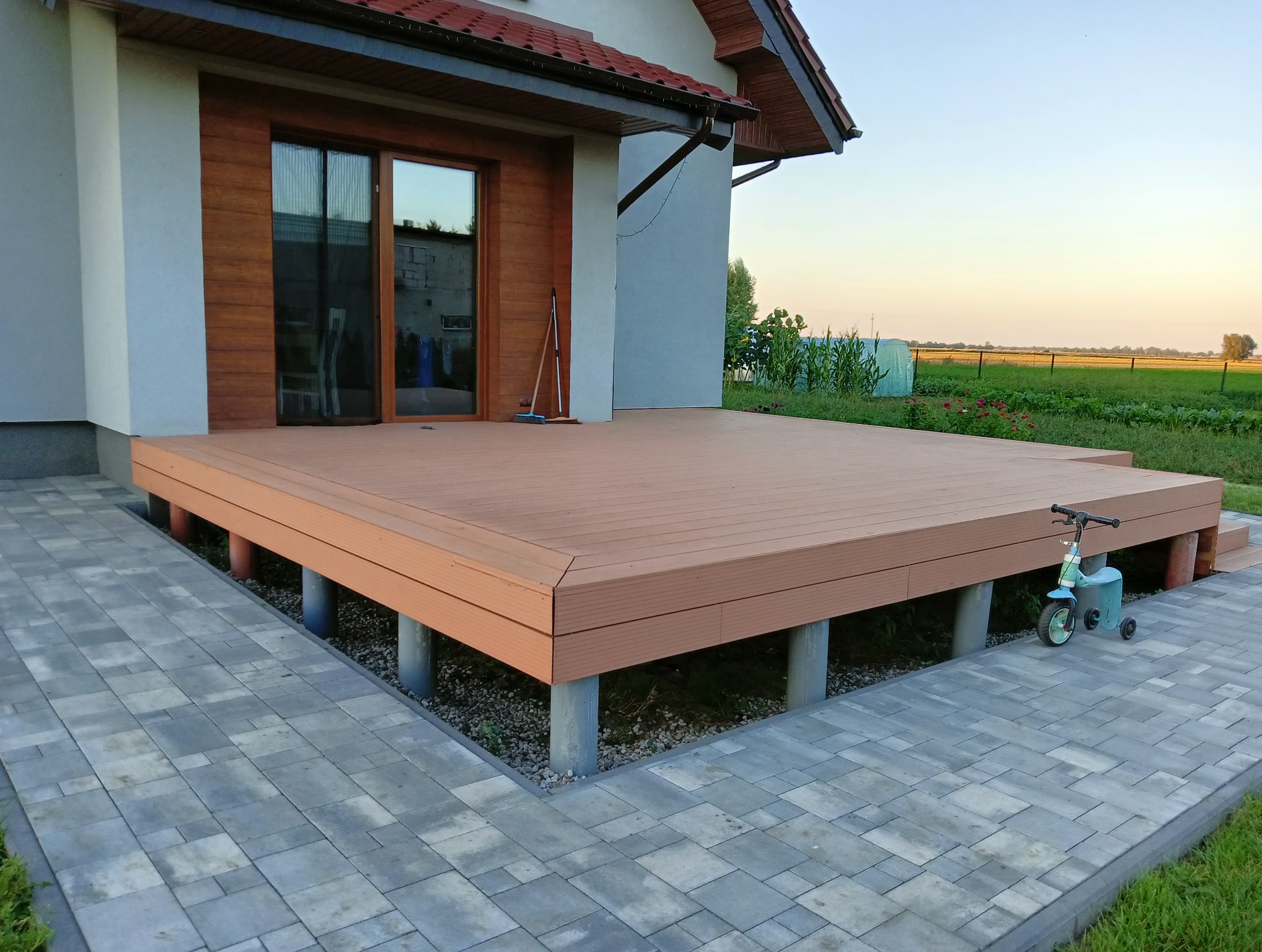 Nowoczesny taras z kompozytu na betonowych słupach, przylegający do domu z przesuwnymi drzwiami. W tle pole i błękitne niebo o zmierzchu. Obok stoi dziecięca hulajnoga.