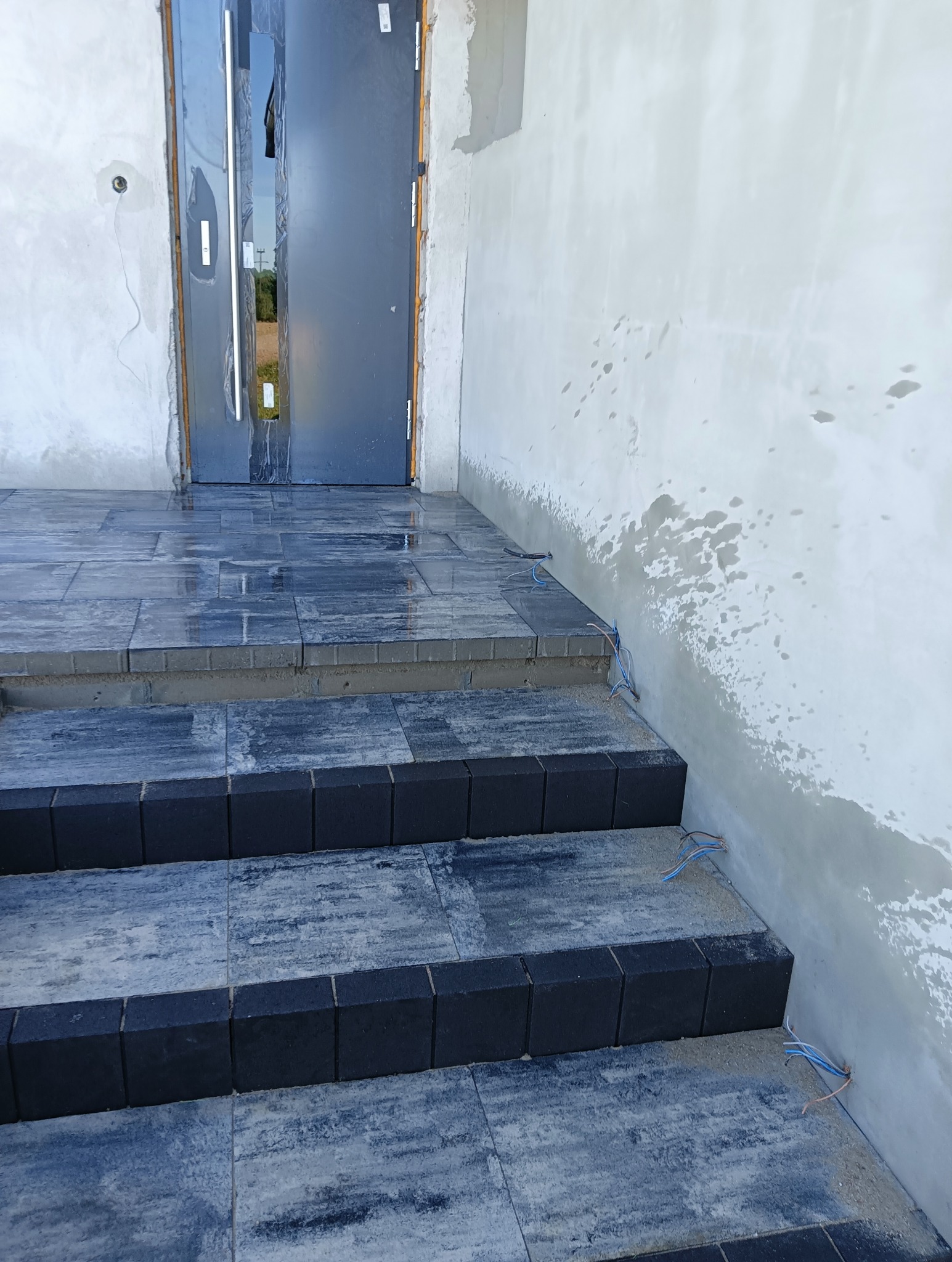 Wykończone schody wejściowe z szarej kostki brukowej z czarnymi obrzeżami, prowadzące do drzwi wejściowych. Ściana w trakcie szpachlowania, widoczne wystające przewody.