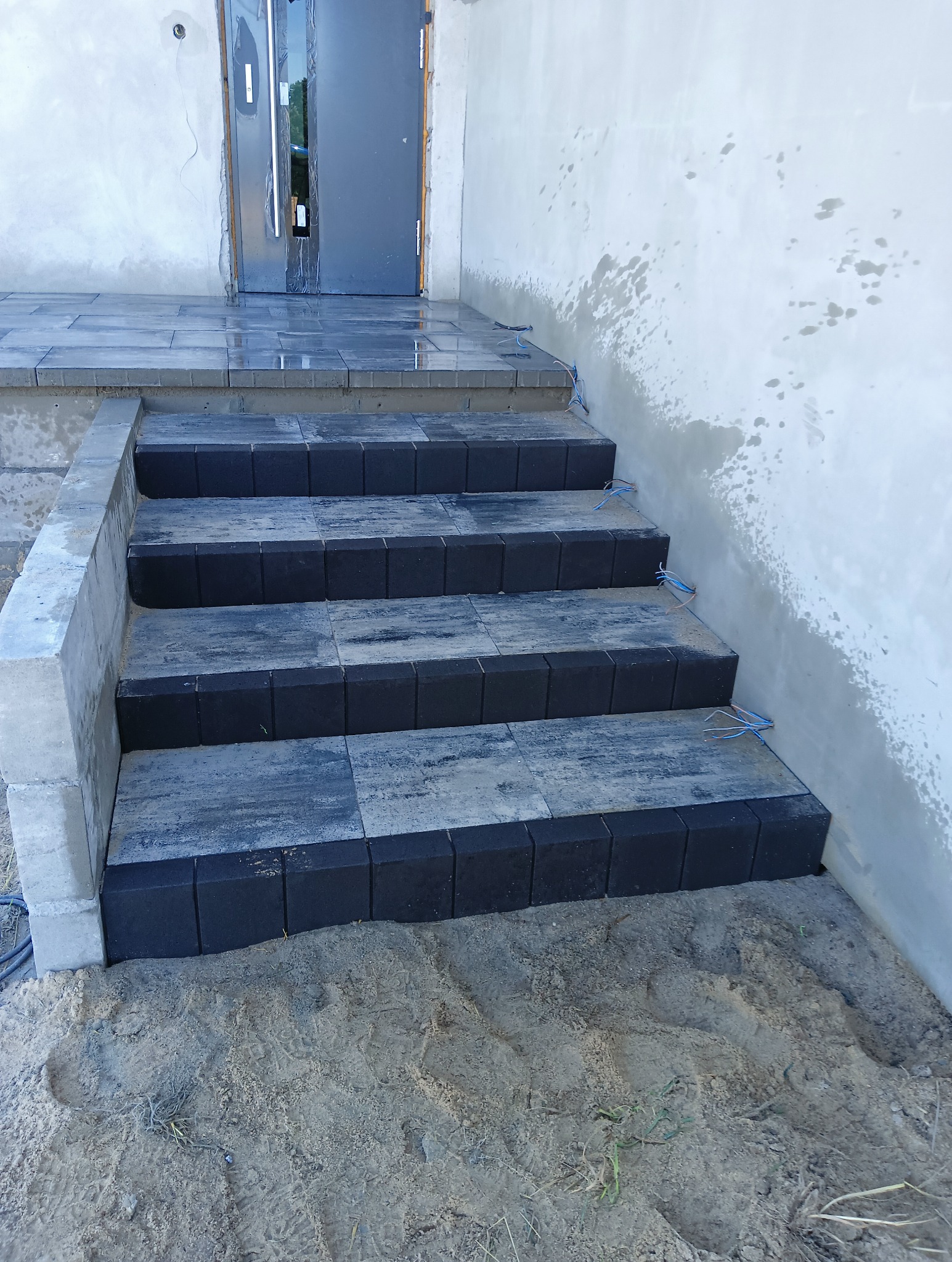 Wykończone schody wejściowe z szarej kostki brukowej z czarnym obrzeżem, prowadzące do drzwi wejściowych. Widoczne niedociągnięcia na ścianie i piasek wokół schodów.