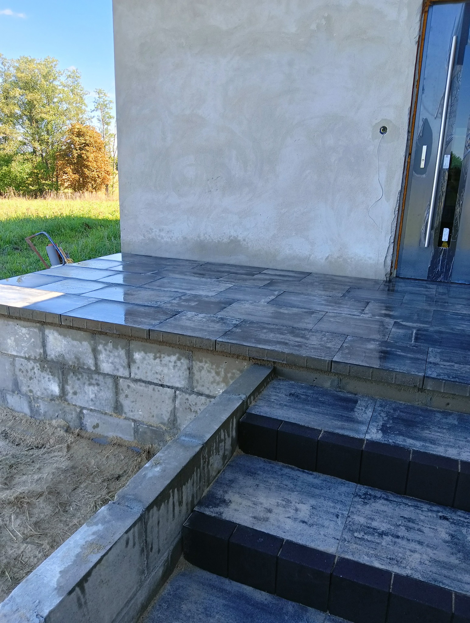 Wykończony taras z kostki brukowej przy wejściu do budynku. Widoczne schody i świeżo położona kostka, na której miejscami widać mokre plamy.