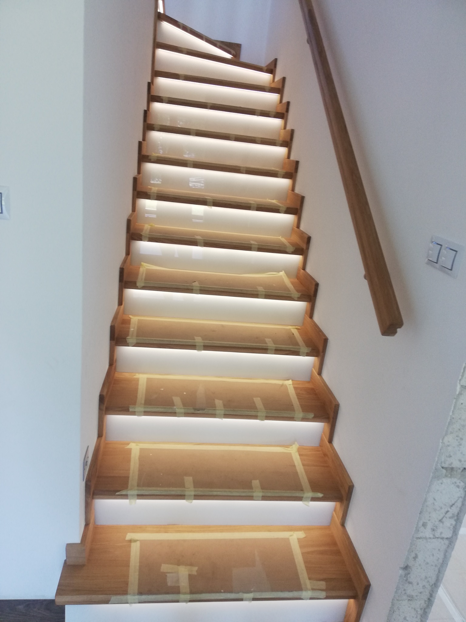 Drewniane schody z podświetleniem LED, zabezpieczone taśmą malarską. Widok z dołu, ukazujący detale wykończenia i oświetlenia. Nowoczesny design.