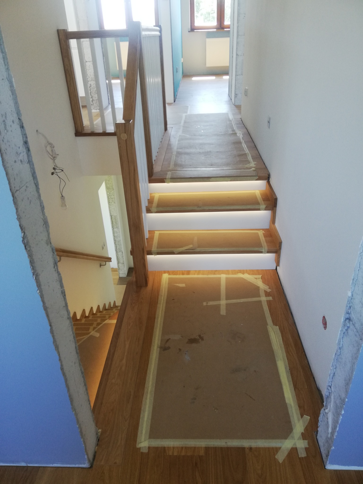 Drewniane schody z podświetleniem LED w trakcie montażu, zabezpieczone folią i taśmą malarską. Widok z góry na klatkę schodową w surowym stanie.
