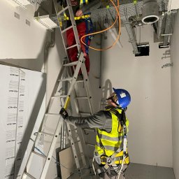 SYSTEMY24 SPÓŁKA Z OGRANICZONĄ ODPOWIEDZIALNOŚCIĄ - Dwóch monterów na drabinie instaluje przewody w budynku. Jeden w kasku i kamizelce a drugi na górze drabiny, widoczne metalowe koryta kablowe i rury.