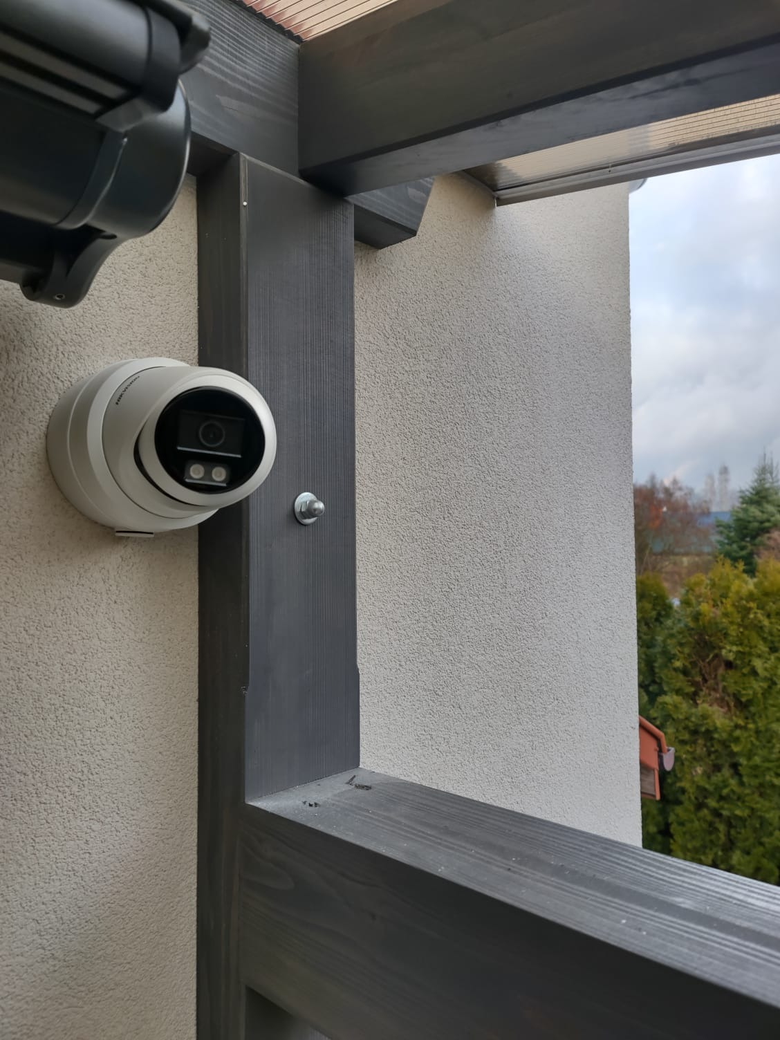 Kamera monitoringu kopułkowa, zamontowana na elewacji budynku z szarą drewnianą konstrukcją. Widok na zewnątrz, drzewa w tle.
