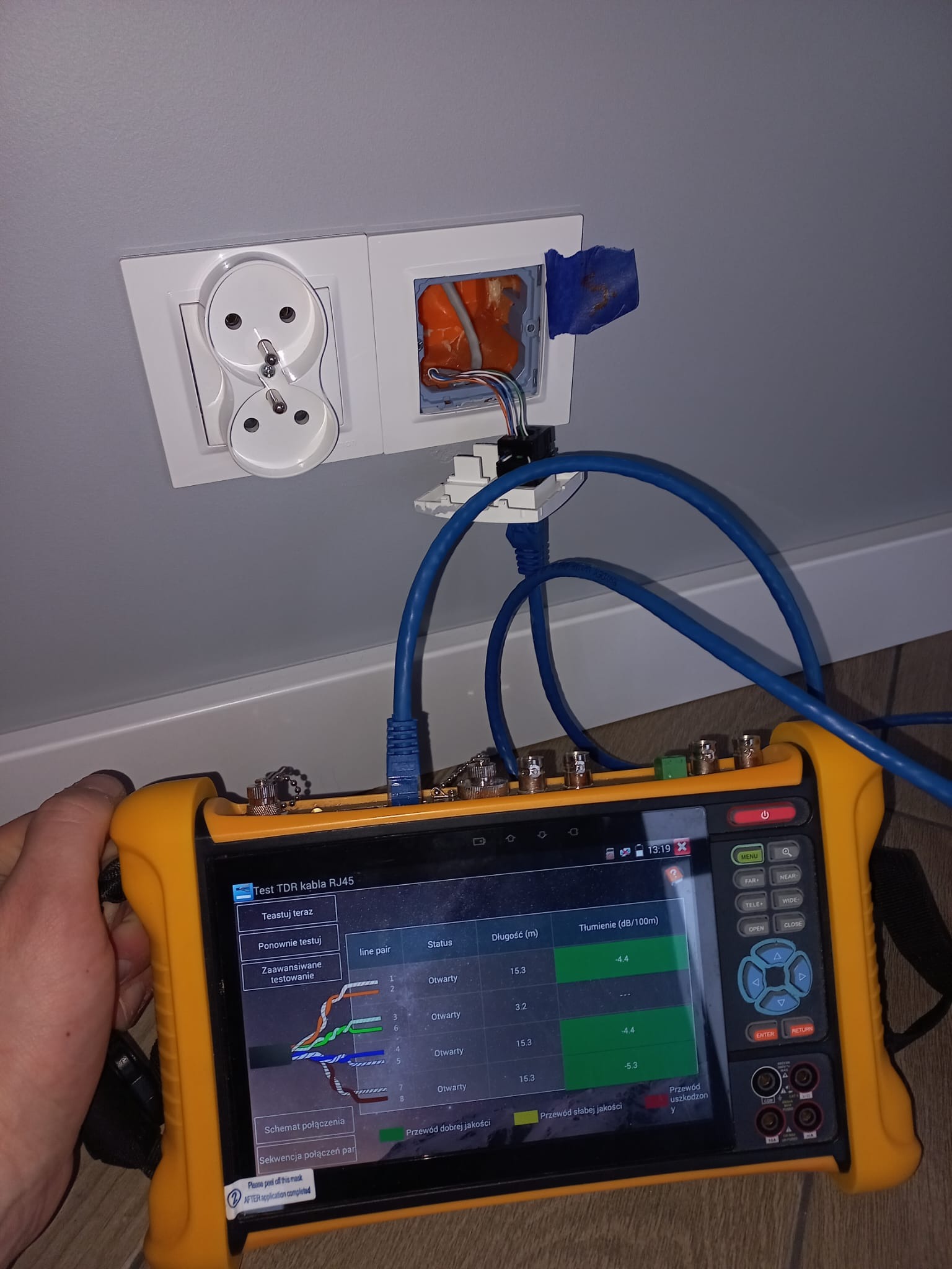 Test kabla RJ45. Tester sieciowy w dłoni, podłączony do gniazda sieciowego w ścianie. Sprawdzanie stanu połączenia, długości kabla i tłumienia sygnału.