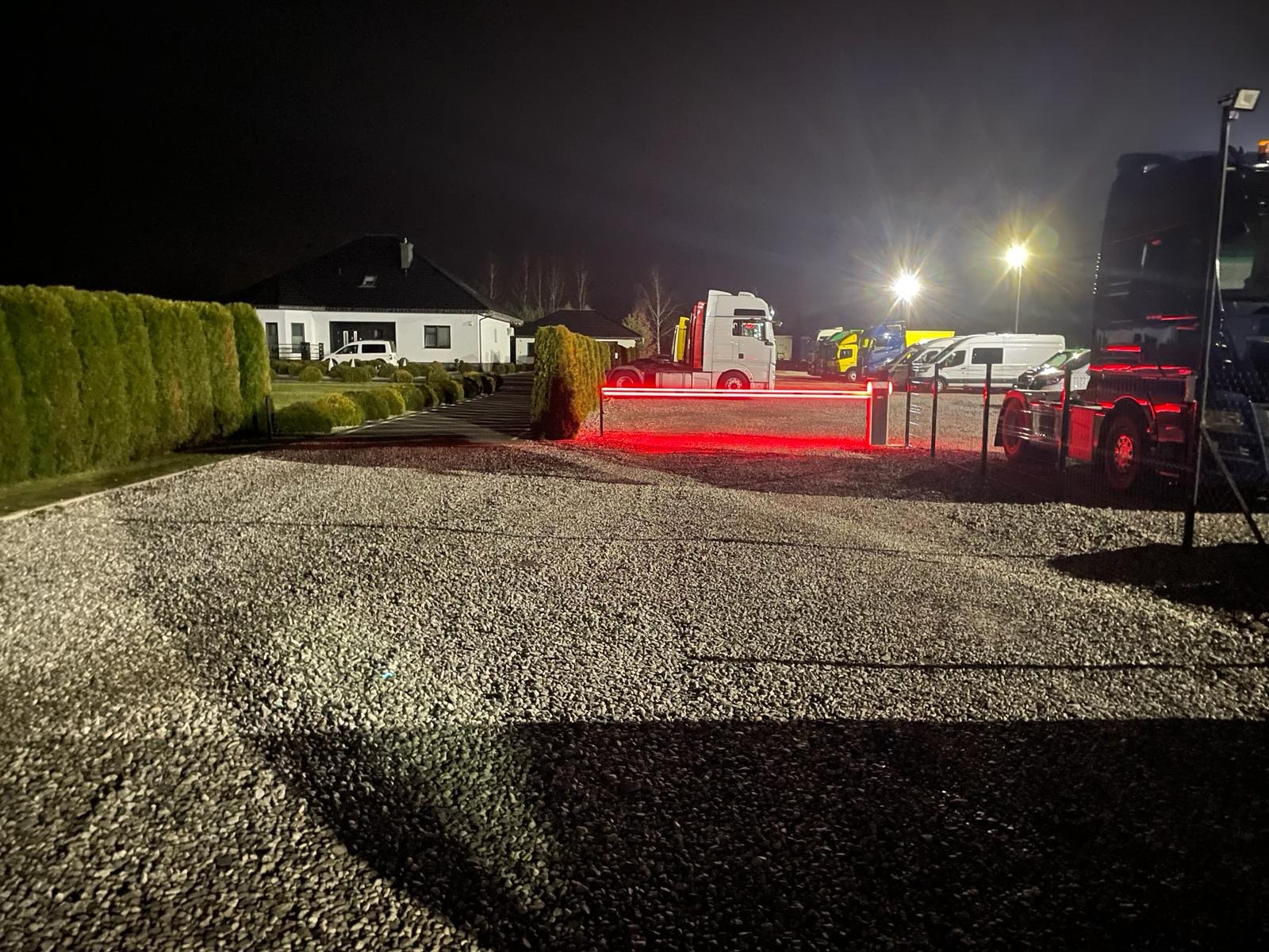 Nocne ujęcie parkingu z automatyczną barierą świetlną, zaparkowanymi ciężarówkami i oświetlonym budynkiem w tle. Widoczna żwirowa nawierzchnia.