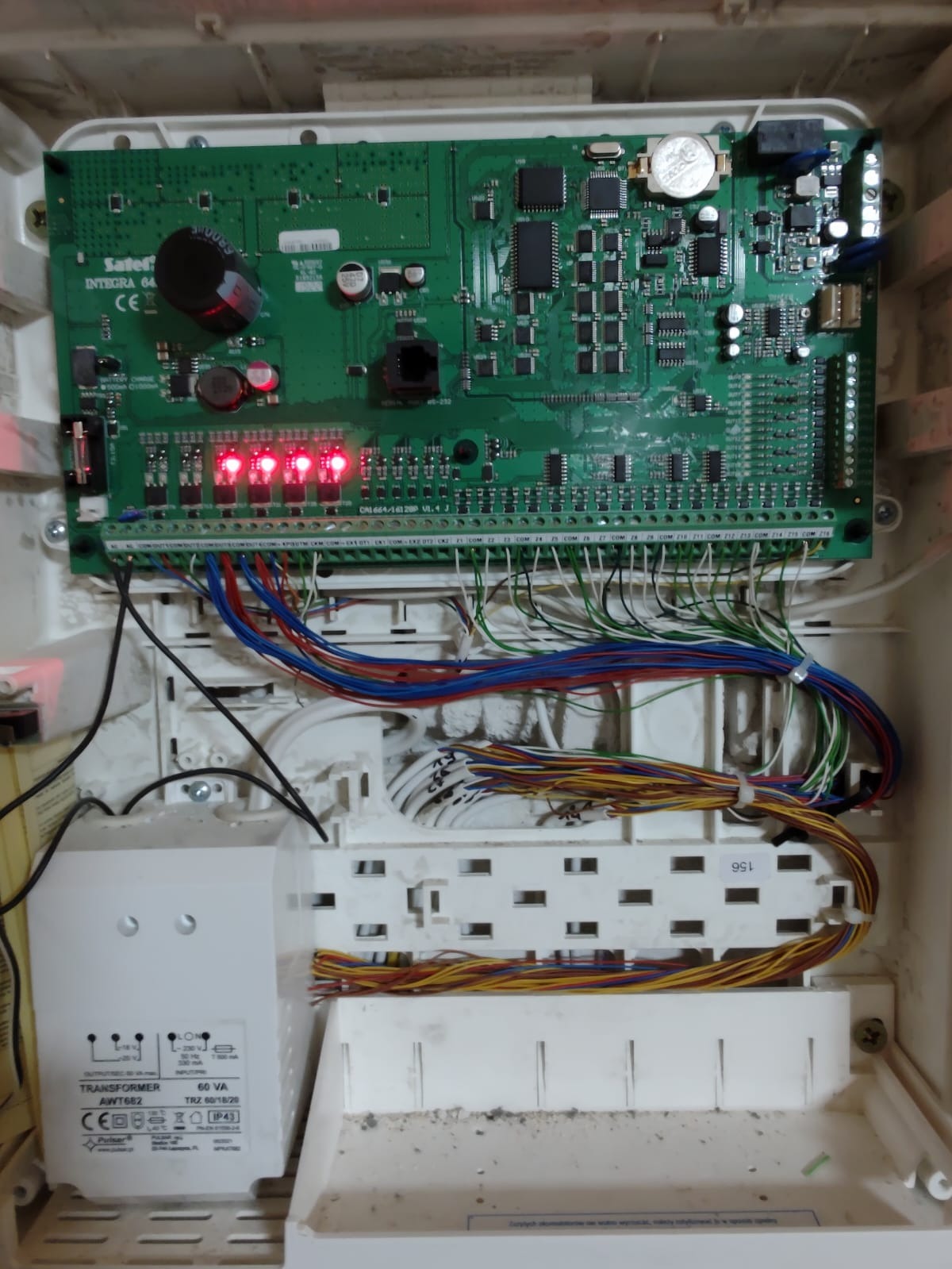 Otwarta obudowa centrali alarmowej Satel Integra 64 z widoczną elektroniką, licznymi przewodami i transformatorem. Zapalone diody sygnalizacyjne.