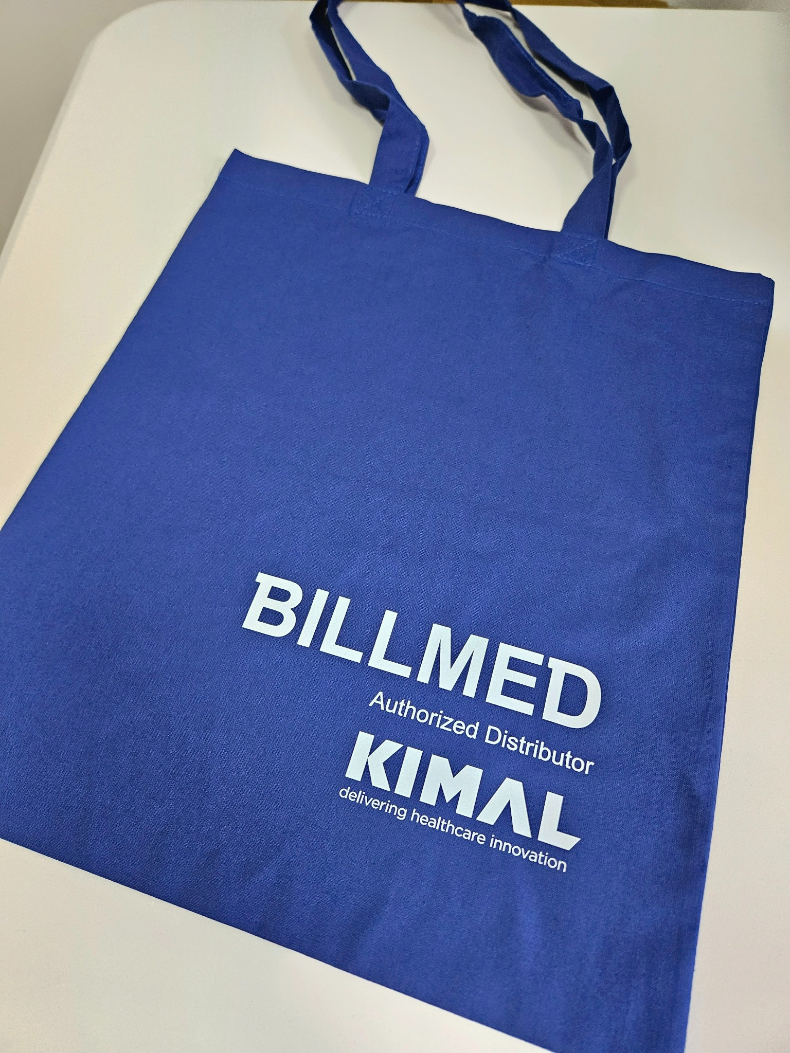 Niebieska torba z nadrukiem 'BILLMED Authorized Distributor KIMAl delivering healthcare innovation'. Torba leży na białym tle.