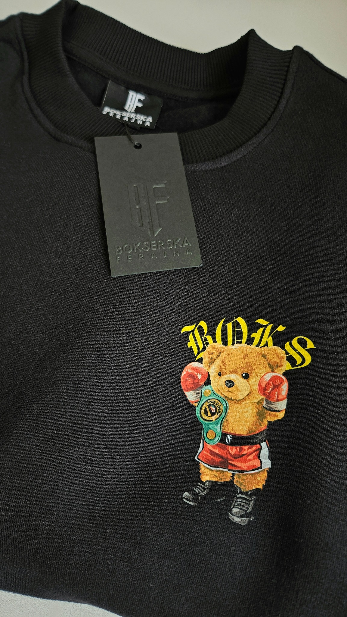 Czarna bluza z nadrukiem misia-boksera na froncie, widoczna metka marki. Detal odzieży z oryginalnym wzorem, prezentujący unikalny projekt graficzny.