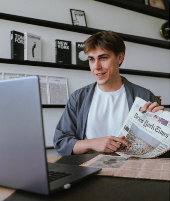 Młody mężczyzna z gazetą 'New York Times' prowadzi lekcję online na laptopie. W tle książki 'London', 'Paris' i grafika.