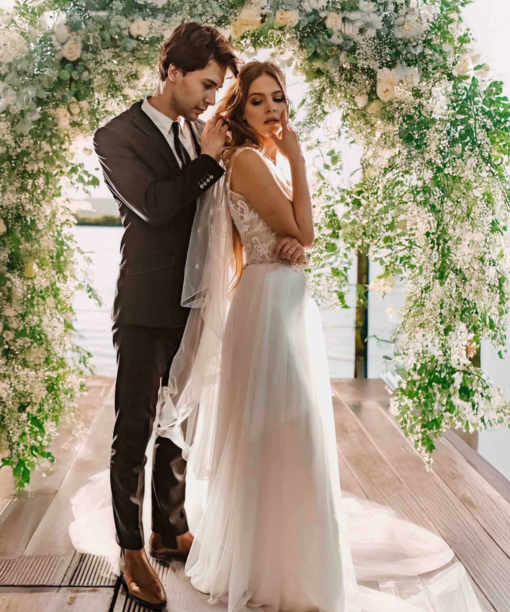 Para młoda pod kwiatowym łukiem. Pan młody poprawia welon pannie młodej. Jasne, naturalne światło, drewniana podłoga, delikatne kwiaty.