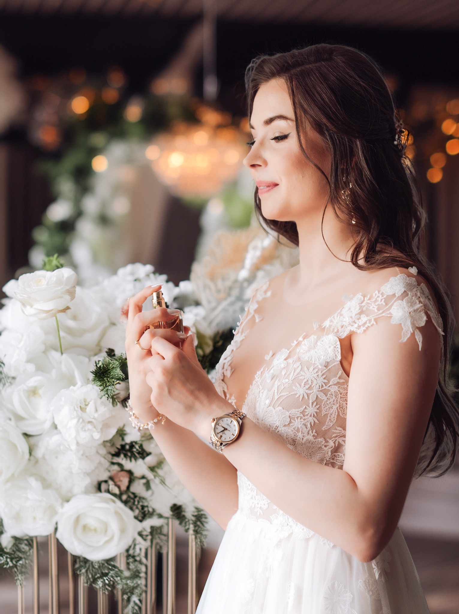 Panna młoda w koronkowej sukni spryskuje się perfumami przed ślubem, na tle białych róż i rozmytych światełek.