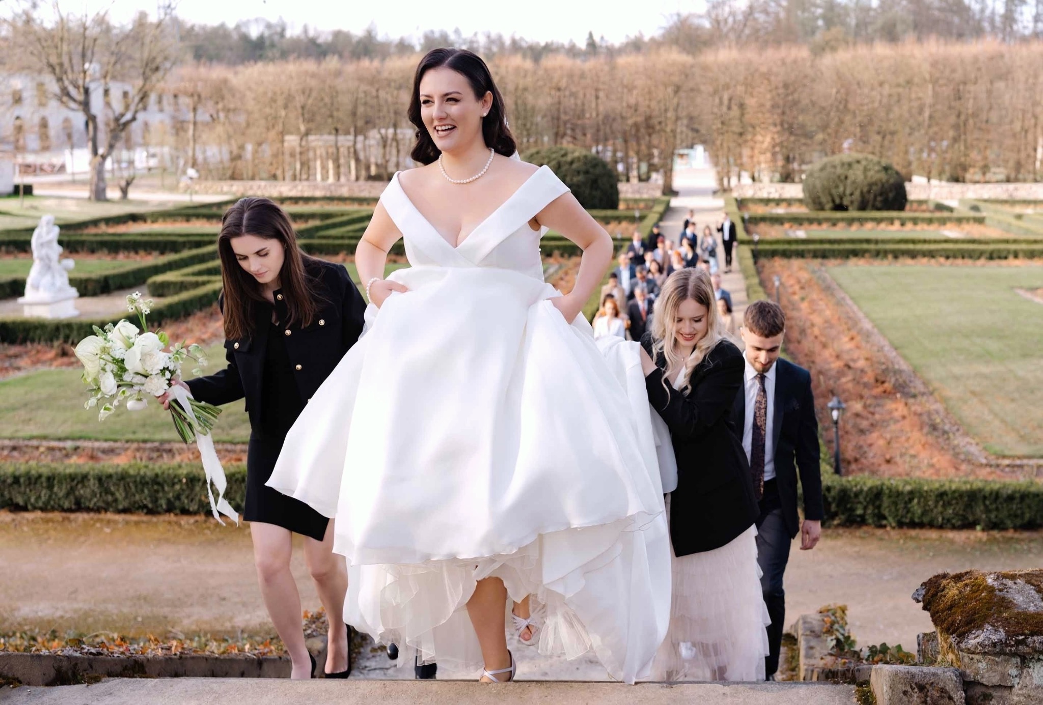 Uśmiechnięta panna młoda w białej sukni schodzi po schodach w parku, w tle goście weselni i klasycystyczny pałac. Jasne, naturalne światło.