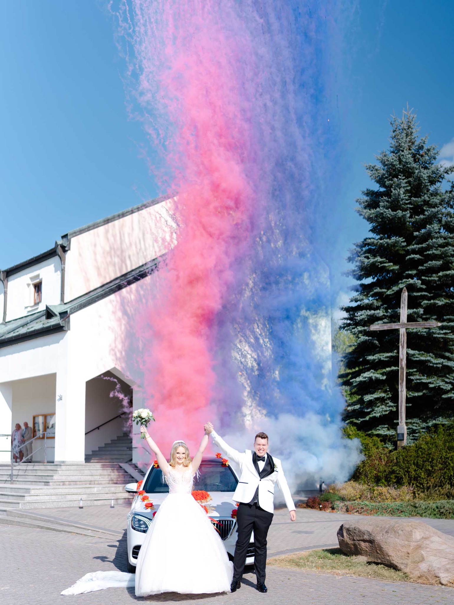 Młoda para świętuje z kolorowym dymem przed udekorowanym samochodem i kościołem w tle, trzymając się za ręce.
