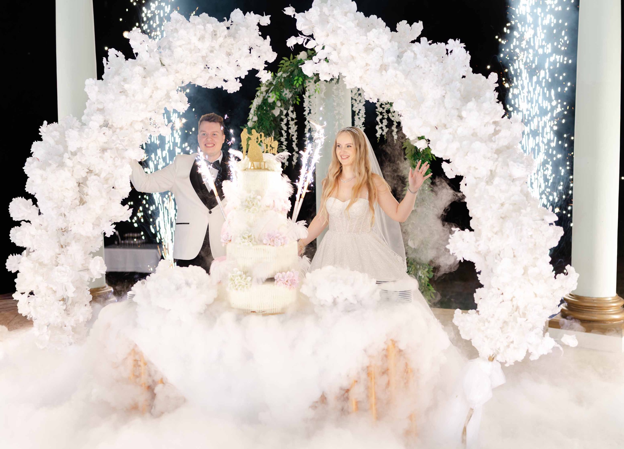 Para młoda w Warszawie świętuje wesele pod dekoracyjnym łukiem z kwiatów i fontannami iskier, z tortem. Uśmiechnięci, w eleganckich strojach.