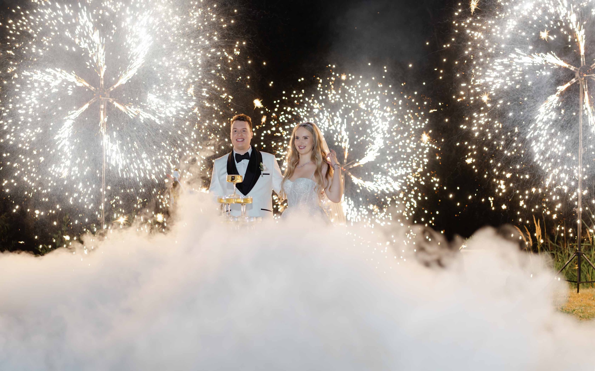 Para młoda w dymie i blasku fontanny iskier, świętuje z tortem z szampana. Uśmiechnięci, w eleganckich strojach ślubnych, na tle nocnego nieba.