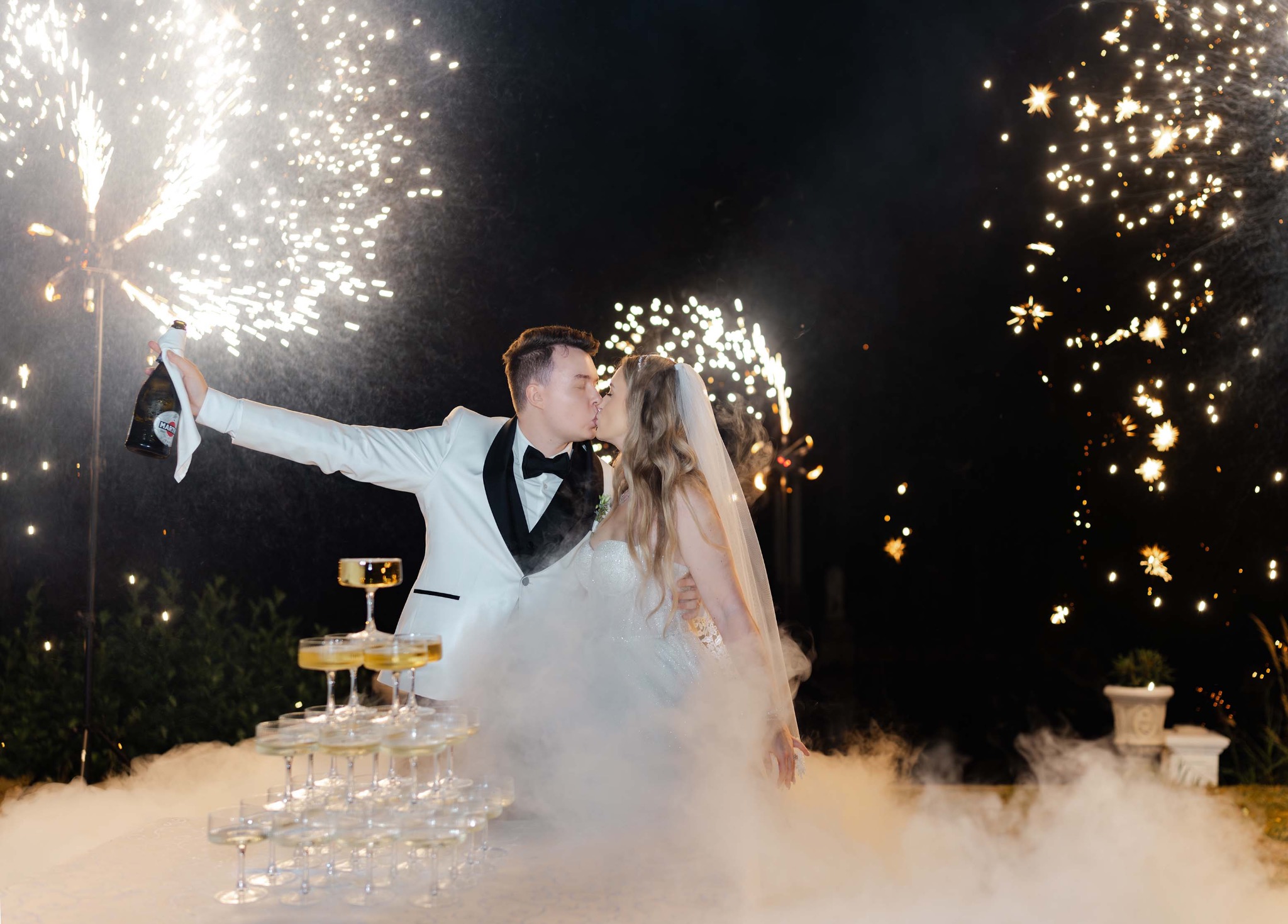 Pocałunek pary młodej na tle fontanny iskier i piramidy z kieliszków, groom w białym smokingu unosi butelkę Martini. Romantyczna atmosfera.