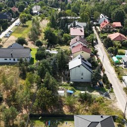 Flyxpress.pl - Filmy z Drona Warszawa - Widok z drona na osiedle domów jednorodzinnych w słoneczny dzień. Domy z różnymi kolorami dachów otoczone zielenią drzew. W tle widoczna droga i zaparkowane samochody.