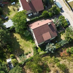 Flyxpress.pl - Filmy z Drona Warszawa - Zdjęcie z drona: dom z czerwoną dachówką, zielony trawnik, drzewa i droga. Ujęcie z góry ukazuje budynek w otoczeniu zieleni i sąsiedniej zabudowy.
