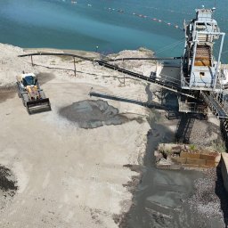 Flyxpress.pl - Filmy z Drona Warszawa - Widok z drona na kopalnię piasku: ładowarka kołowa, taśmociąg i zbiornik wodny.  Krajobraz przemysłowy z błękitną wodą w tle.