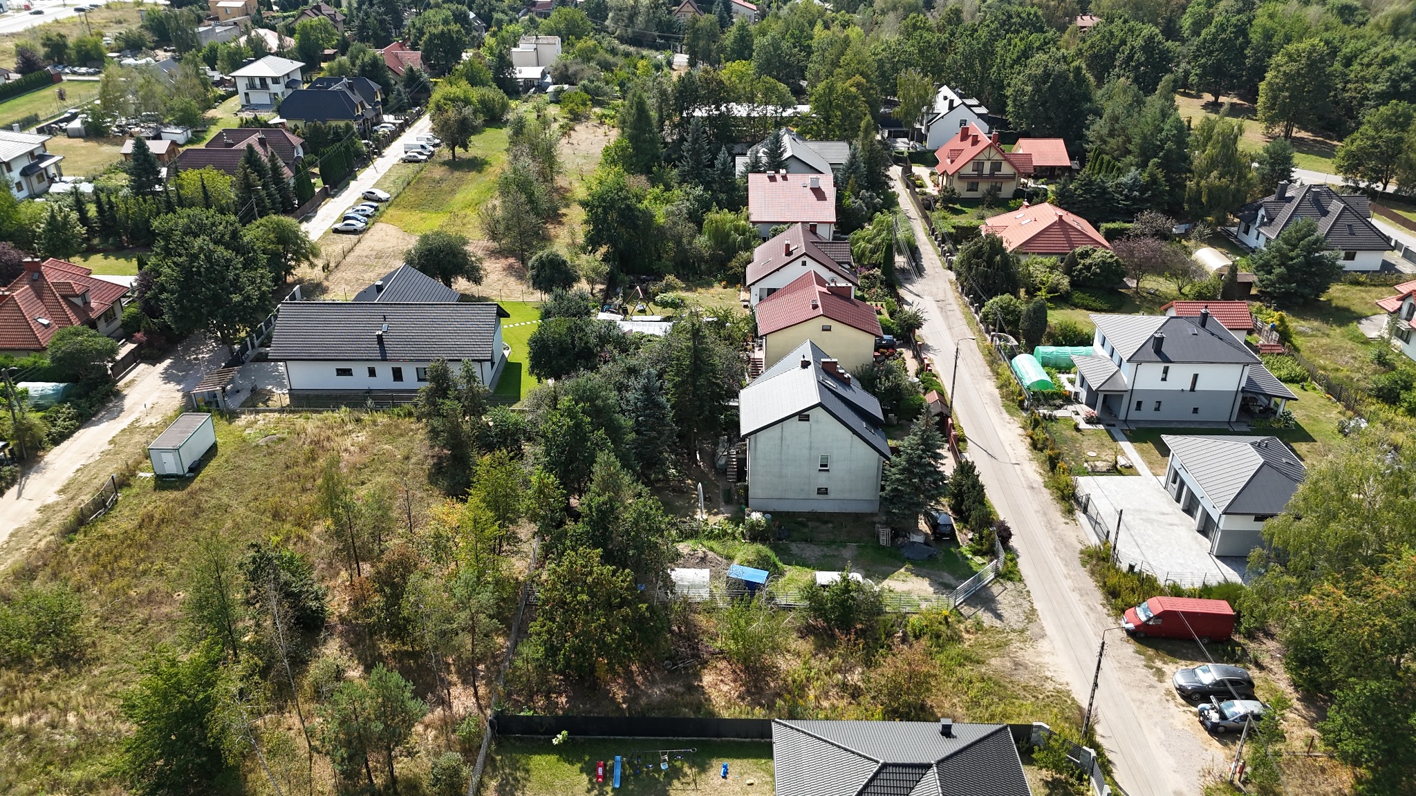 Widok z drona na osiedle domów jednorodzinnych w słoneczny dzień. Domy z różnymi kolorami dachów otoczone zielenią drzew. W tle widoczna droga i zaparkowane samochody.