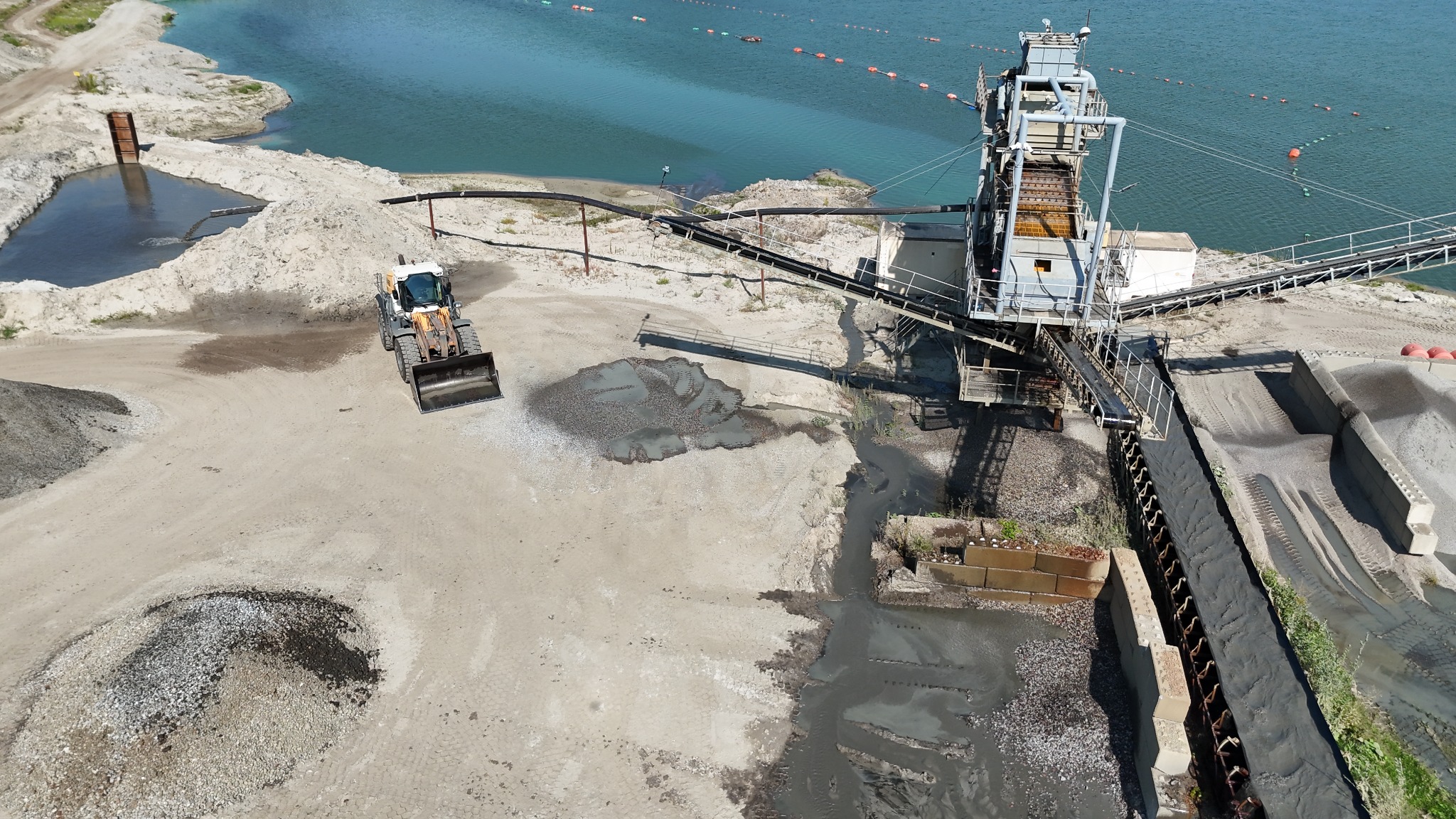 Widok z drona na kopalnię piasku: ładowarka kołowa, taśmociąg i zbiornik wodny.  Krajobraz przemysłowy z błękitną wodą w tle.