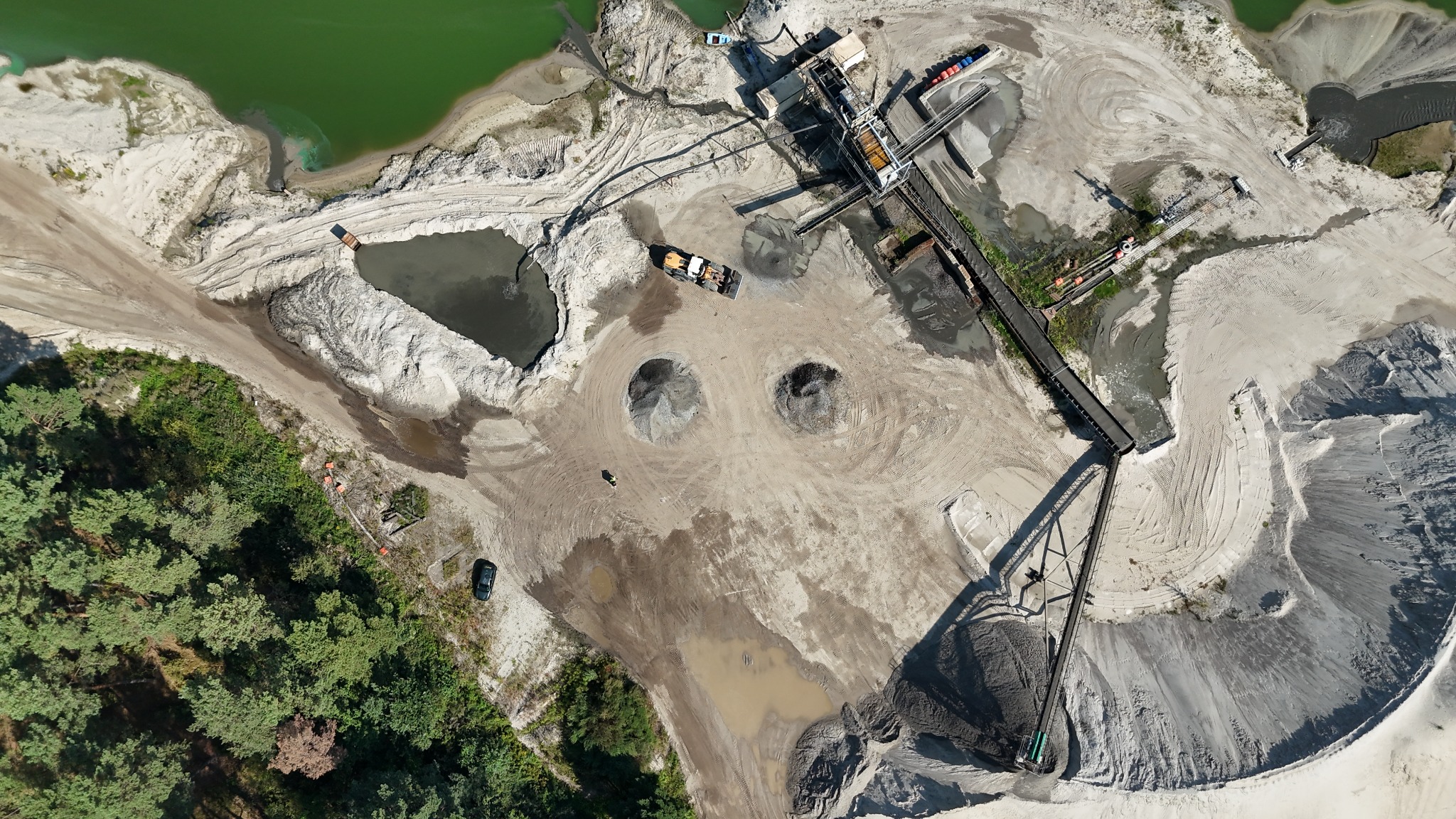Widok z lotu ptaka na kopalnię piasku z zielonym jeziorem, hałdami, sprzętem i drzewami. Długi taśmociąg transportuje urobek. Ślady opon na piasku, maszyny w pracy.