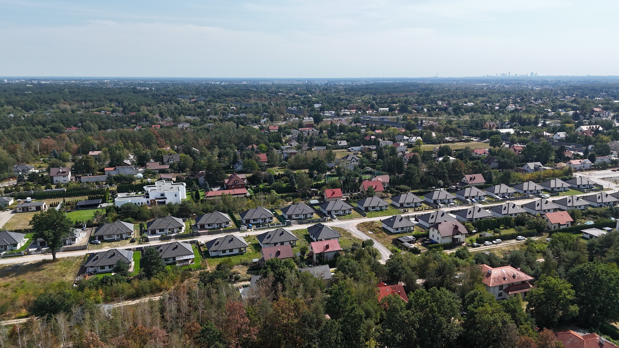 Panoramiczne ujęcie z drona na osiedle domów jednorodzinnych z ciemnymi dachami, otoczone zielenią drzew, z widoczną panoramą miasta w oddali.