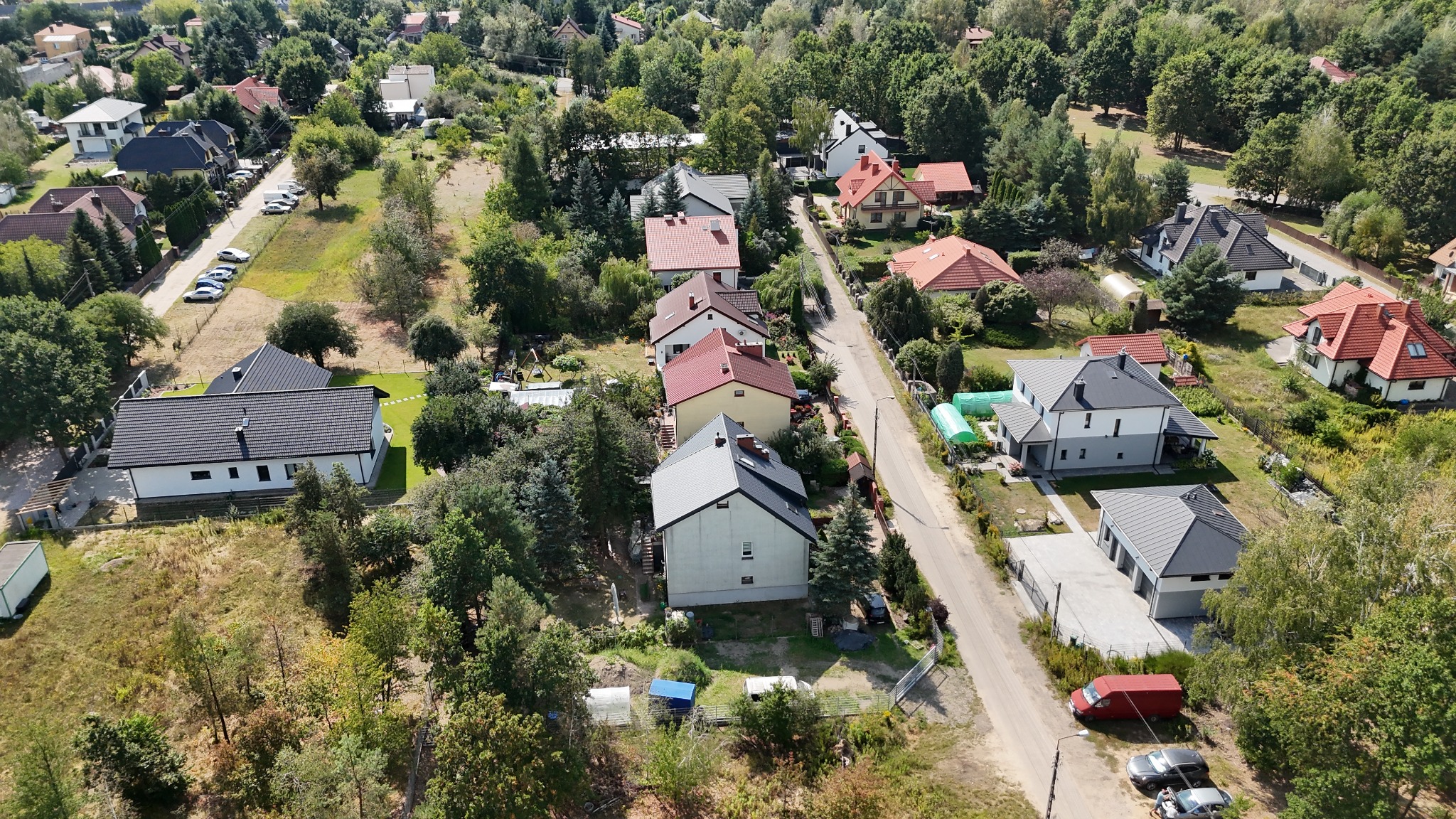 Panoramiczny widok z drona na osiedle domów jednorodzinnych otoczonych zielenią, z widoczną drogą i zaparkowanymi samochodami. Ujęcie w słoneczny dzień.