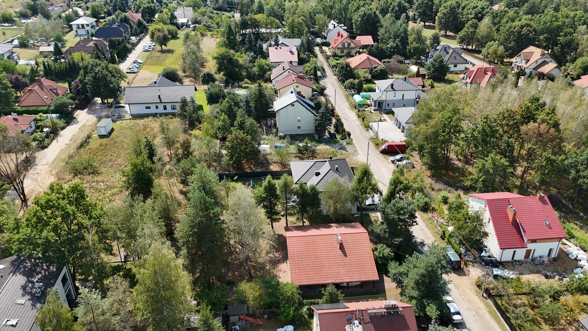 Panoramiczny widok z lotu ptaka na osiedle domów jednorodzinnych z czerwonymi dachami, otoczone zielenią drzew w słoneczny dzień.