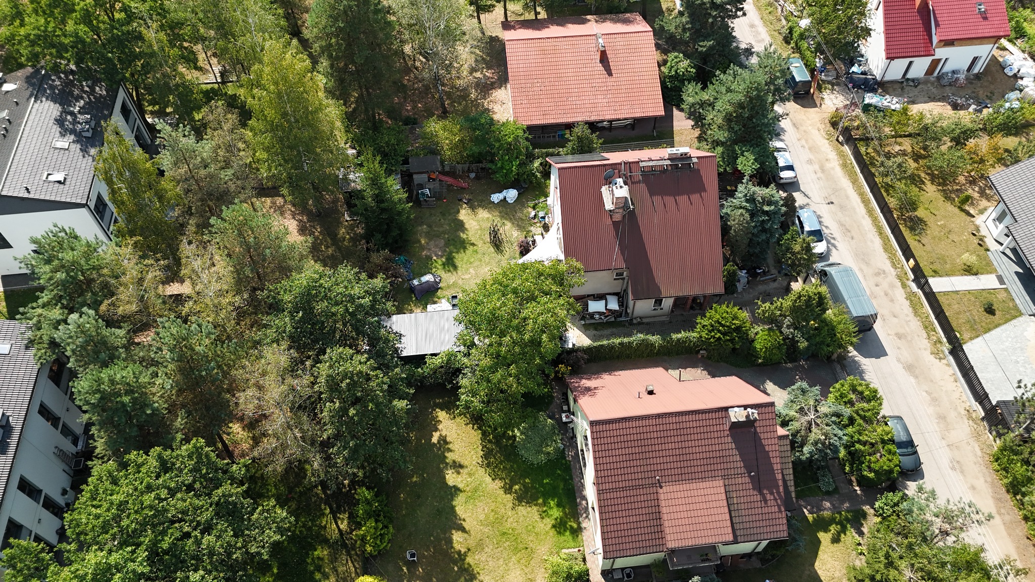 Zdjęcie z lotu ptaka przedstawia osiedle domów jednorodzinnych z czerwonymi dachami, otoczonych zielenią drzew i krzewów, z ulicą i zaparkowanymi samochodami w tle.