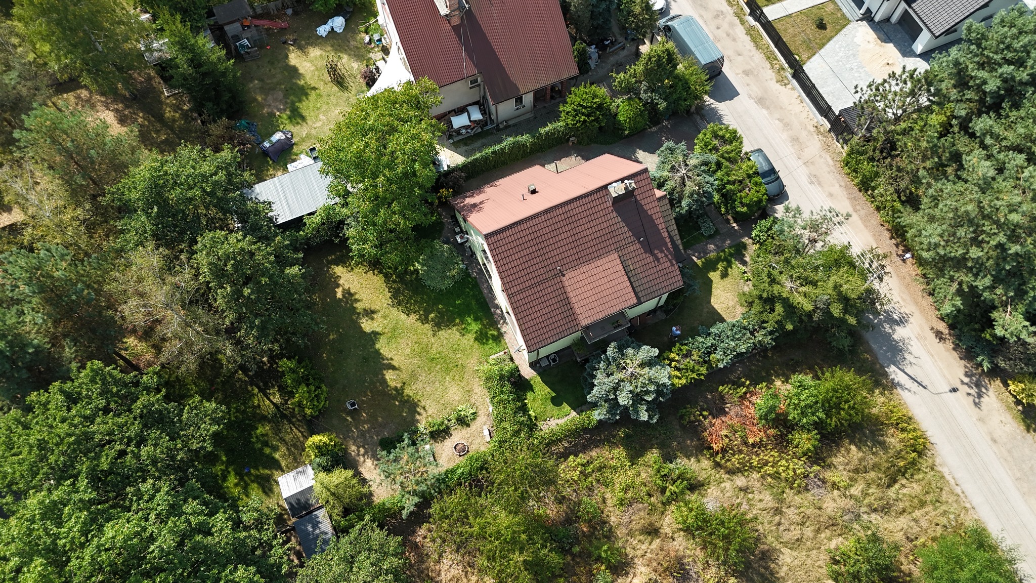 Zdjęcie z drona przedstawia dom z czerwoną dachówką otoczony zielenią, z widoczną drogą i innymi budynkami w tle. Ujęcie z góry, w słoneczny dzień.