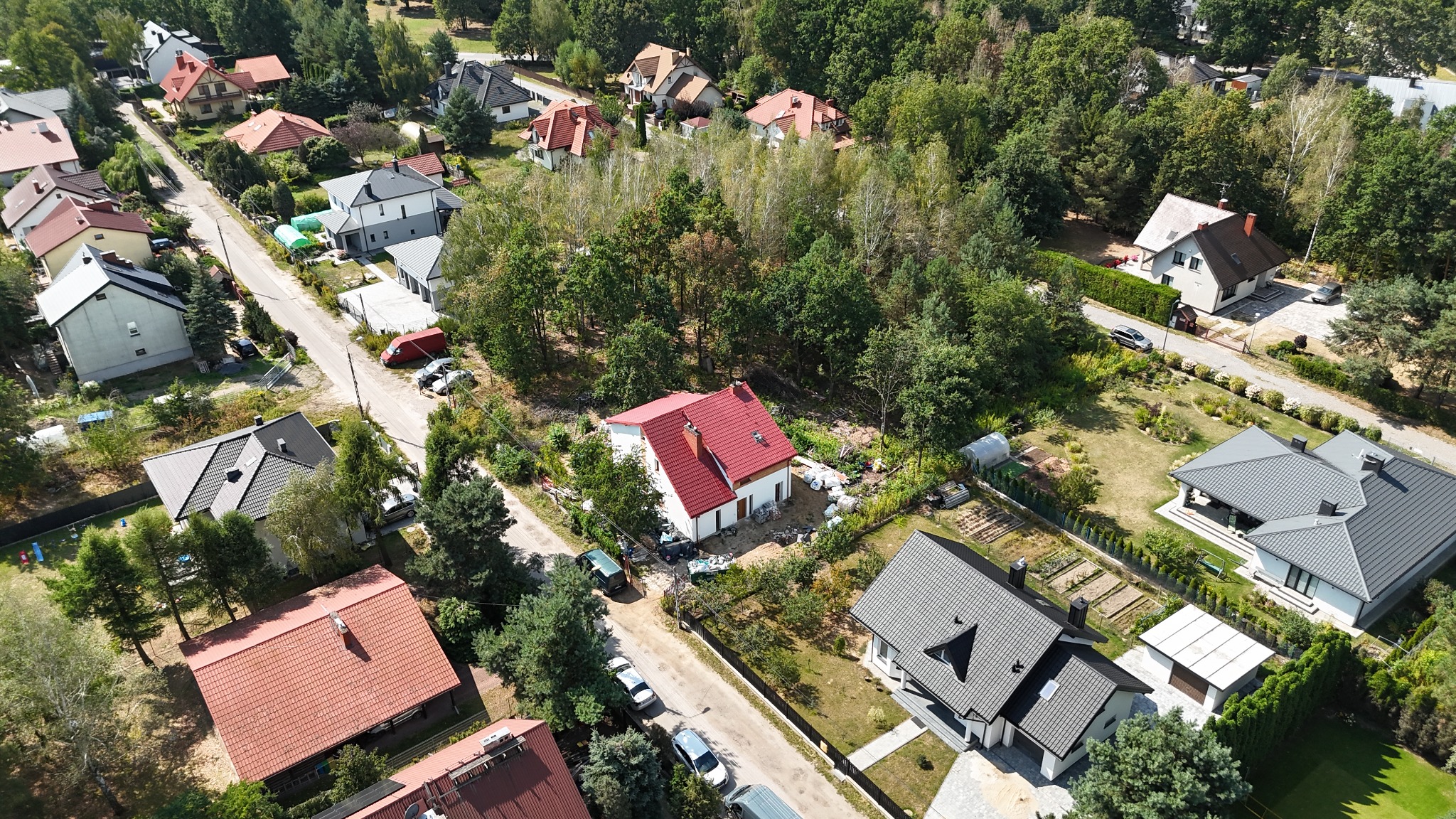 Zdjęcie z drona przedstawia osiedle domów jednorodzinnych z nowym domem z czerwoną dachówką w centrum kadru. Wokół zieleń drzew i krzewów, widoczne samochody na ulicy.
