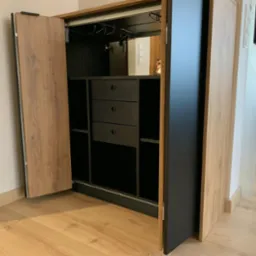 Otwarta szafa wnękowa z drewnianymi drzwiami i czarnym wnętrzem, w kt&oacute;rym widoczne są p&oacute;łki, szuflady i wieszaki na kieliszki. Szafa stoi na drewnianej podłodze.