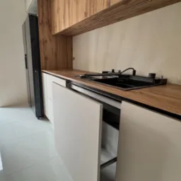 Nowoczesna kuchnia z drewnianymi szafkami i białymi frontami. Widoczny zlew i fragment lodówki. Minimalistyczny design, jasne wnętrze w trakcie montażu.