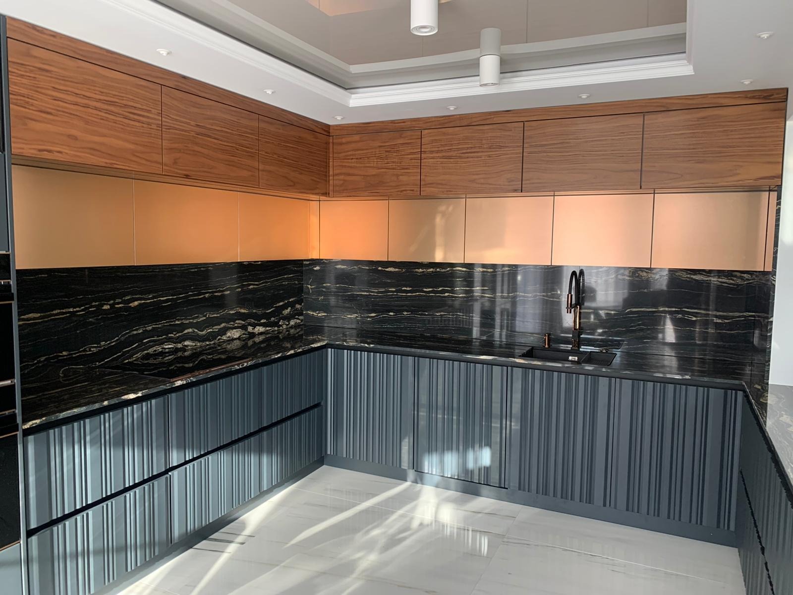 Nowoczesna kuchnia z szarymi, żłobionymi szafkami dolnymi, blatem z czarnego marmuru i górnymi szafkami z drewna orzechowego oraz miedzianymi frontami.
