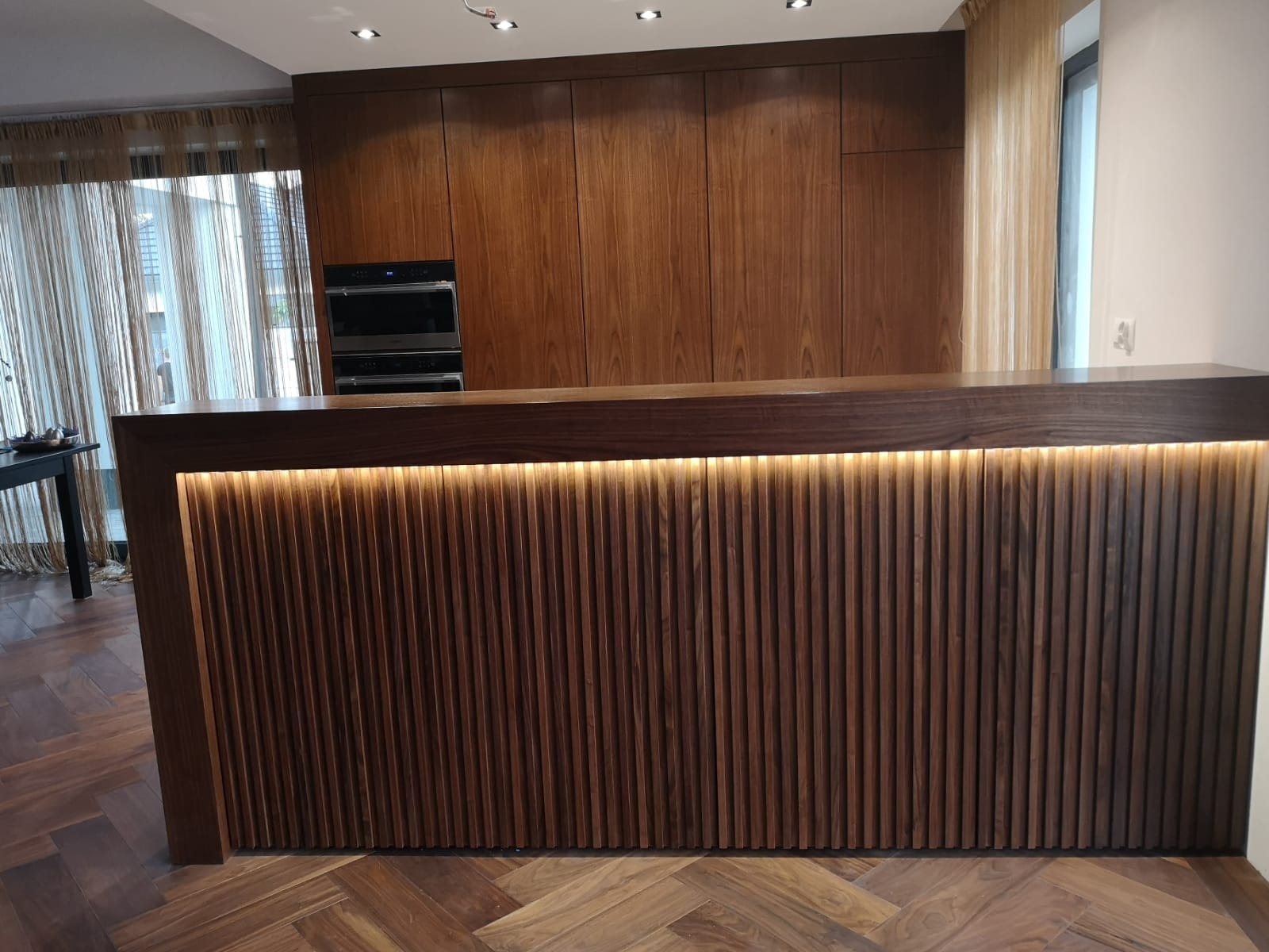 Nowoczesna kuchnia z drewnianą zabudową i oświetlonym barkiem. Parkiet w jodełkę. W tle wbudowany piekarnik i zasłonięte okno.